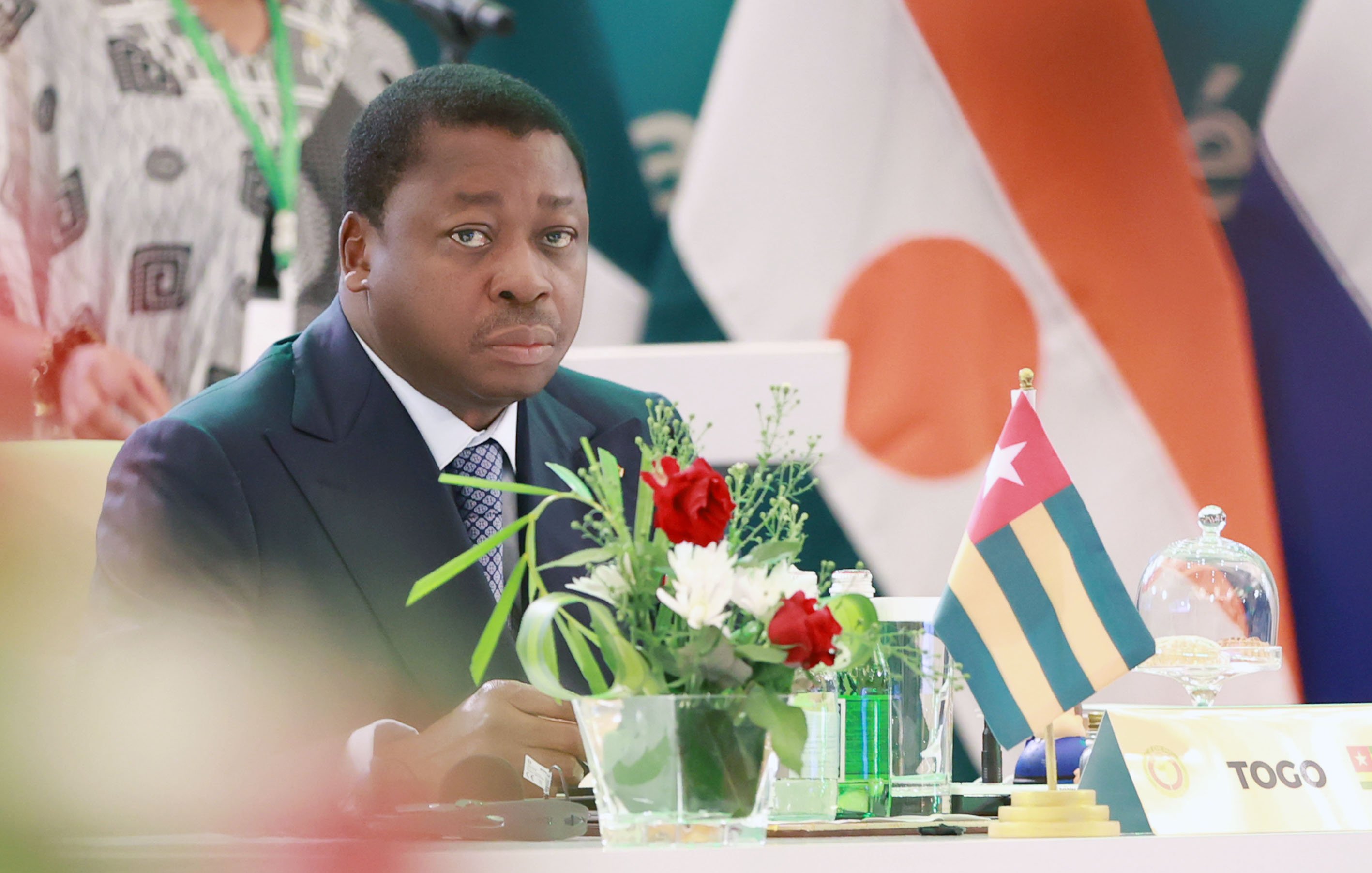 L'image montre un homme assis à une table lors d'un événement officiel. Il porte un costume sombre et une cravate, et son expression semble sérieuse et attentive. Sur la table, il y a un petit vase avec des fleurs colorées, ajoutant une touche de vie à la scène. À côté de lui, on voit un drapeau qui représente le Togo. En arrière-plan, des drapeaux d'autres pays sont également visibles, indiquant un cadre international. L'atmosphère semble formelle et concentrée.