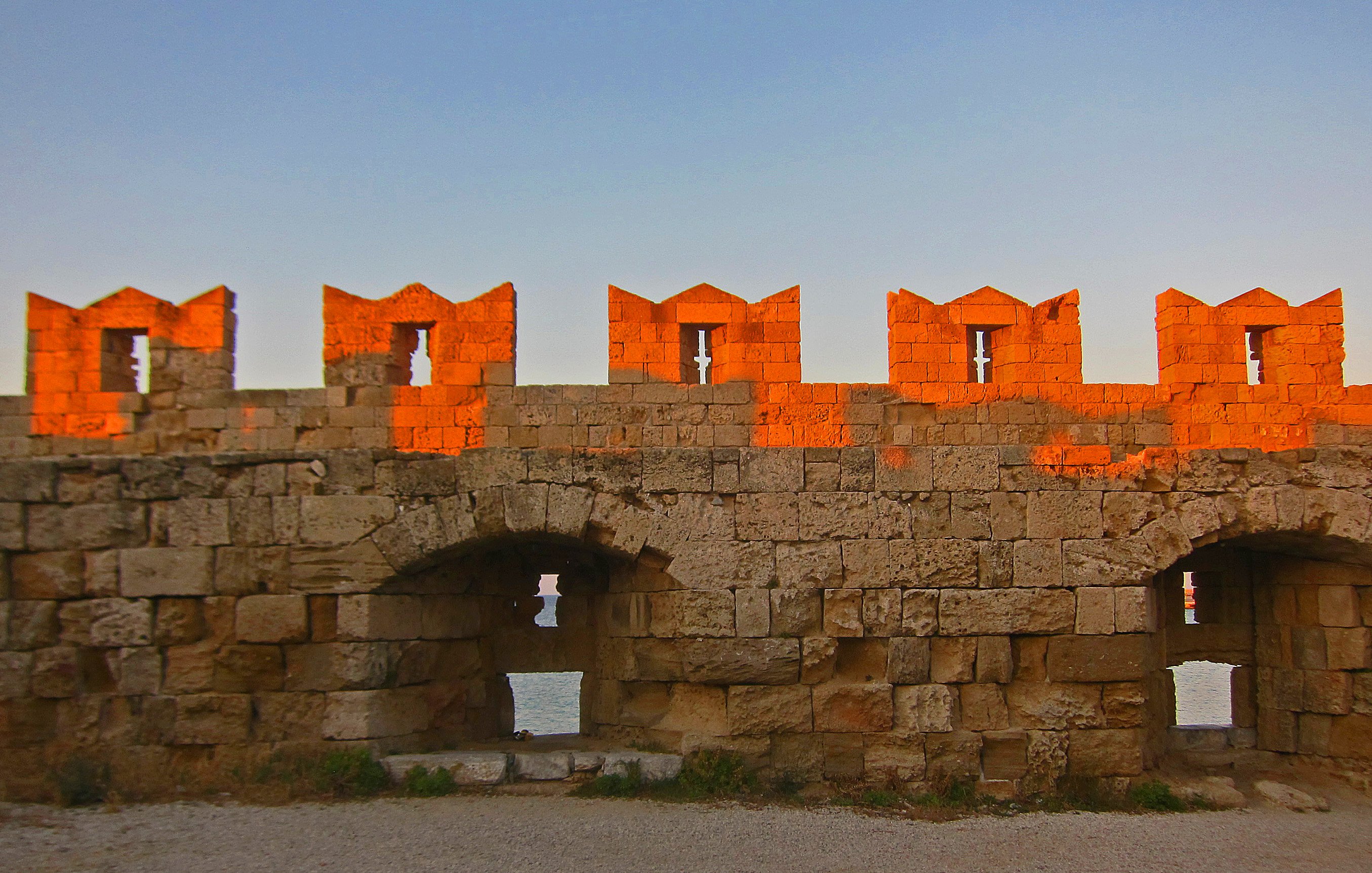 L'image montre un mur de fortification ancien, construit en pierres de taille. La lumière dorée du soleil se reflète sur la pierre, créant un effet chaleureux et pittoresque. Au sommet du mur, on peut voir des créneaux, typiques des châteaux, qui forment des formes découpées. Le mur présente des arches et des ouvertures qui laissent entrevoir un espace en arrière-plan, probablement une vue sur la mer ou un paysage naturel. Le sol est recouvert de gravier, ajoutant une texture à l'environnement. L'ensemble dégage une atmosphère historique et paisible.