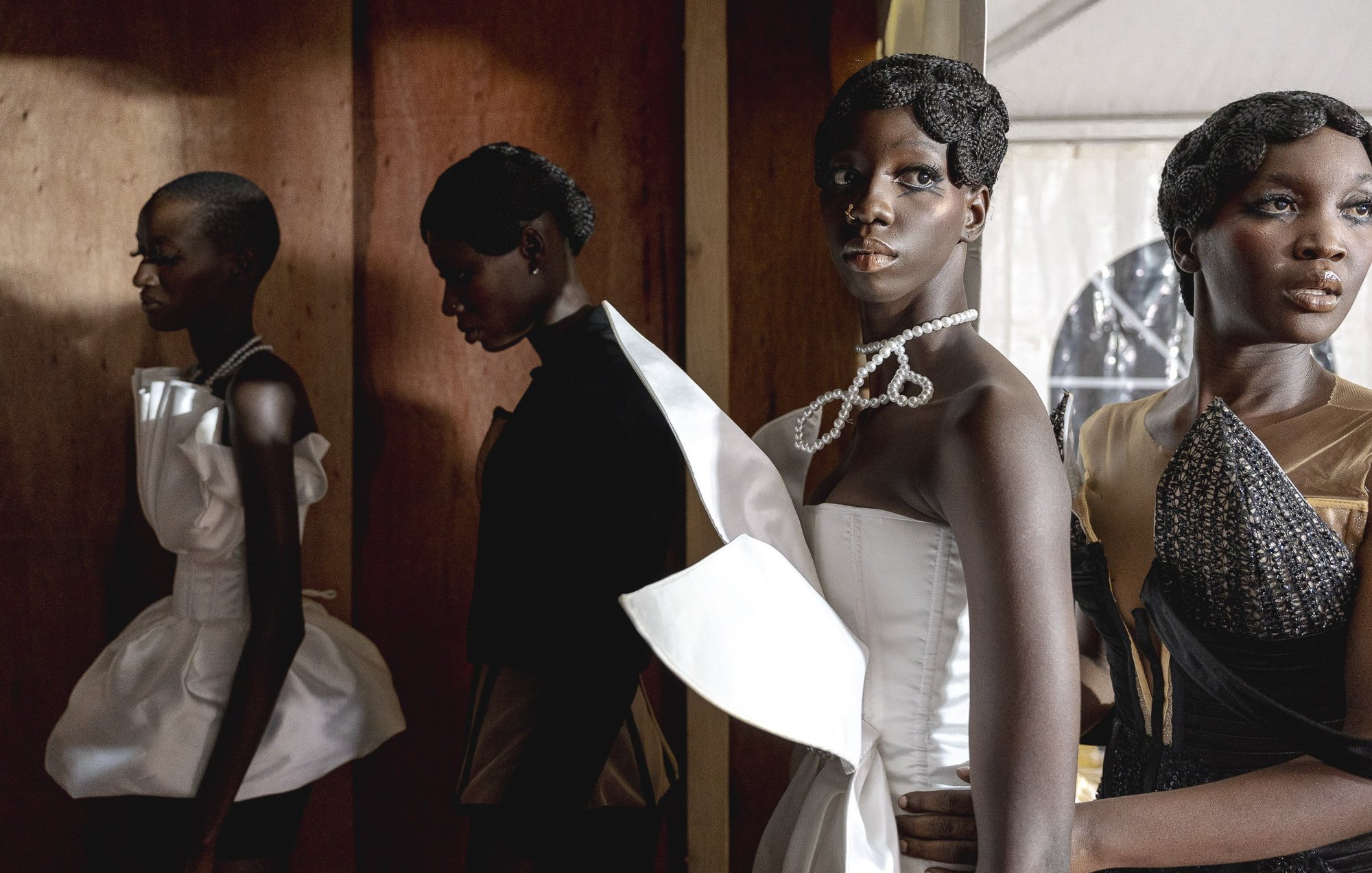 L'image présente un groupe de mannequins lors d'un événement de mode. Les mannequins, d'origines diverses, portent des tenues élégantes et extravagantes, mettant en valeur des tissus texturés et des coupes audacieuses. Au premier plan, l'un des mannequins se distingue par une robe blanche ornée d'un grand nœud, tandis que les autres vêtus de créations plus sombres, affichent des coiffures soignées. La lumière joue sur les tissus, créant un contraste entre les ombres et les zones éclairées, ce qui ajoute une profondeur visuelle à la scène. L'ambiance est à la fois chic et artistique, capturant l'énergie dynamique du monde de la mode.