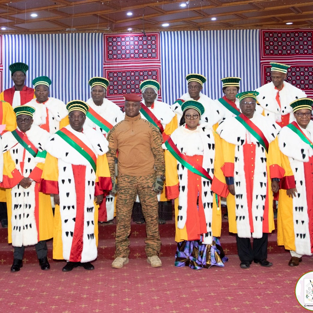 The image features a formal gathering of a group of individuals, likely in a ceremonial setting. They are wearing traditional attire, which includes colorful robes adorned with striped and patterned elements. Many of them are also wearing distinctive hats or headgear. The setting appears to be well-decorated, possibly for an official event, with a red carpet and banners or decorations in the background. A few individuals stand out in military or official uniforms, suggesting a mix of civilian and military presence at the event.