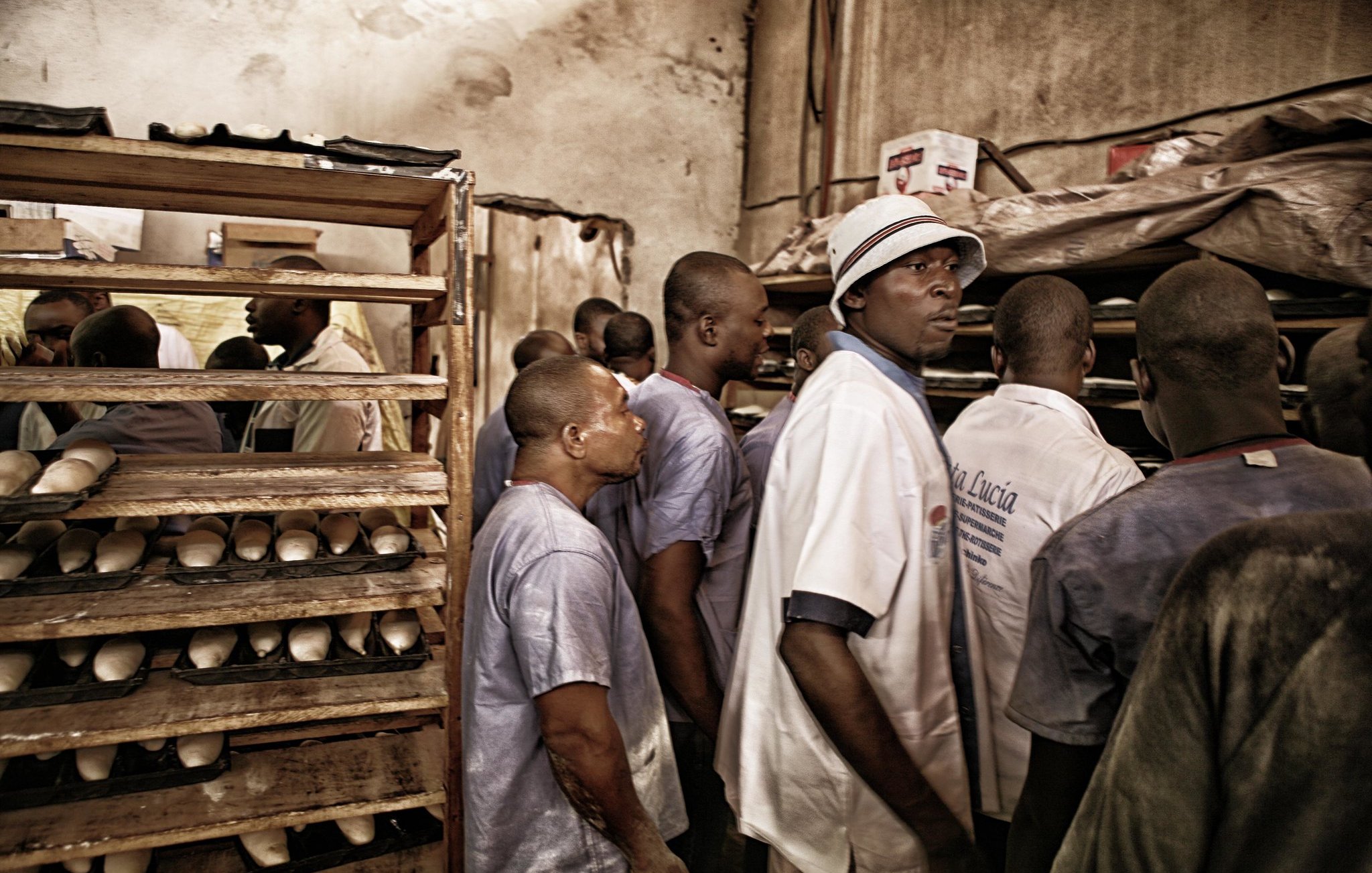Dans cette image, nous voyons un intérieur sombre et chargé, probablement un espace de travail. Plusieurs hommes se tiennent debout, leurs expressions montrent de l'attention ou de la curiosité. Ils portent des vêtements simples, notamment des chemises et des chapeaux, créant une atmosphère de camaraderie. À l'arrière-plan, on aperçoit des étagères en bois remplie de ce qui semble être des objets enroulés ou des produits. L'éclairage est doux, ajoutant une ambiance de chaleur au lieu. L'ensemble évoque un moment de partage ou de travail collectif.