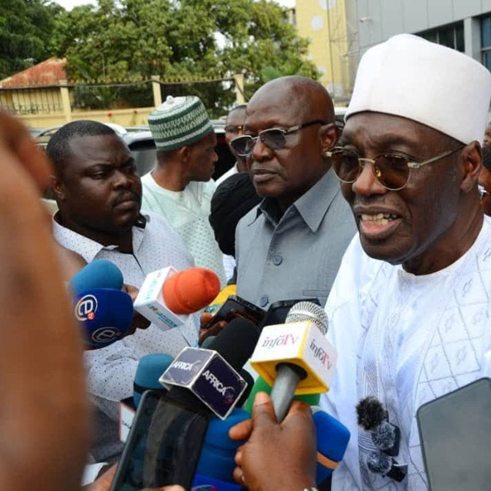 The image shows a group of people gathered around a man wearing a white traditional outfit and a white cap. He appears to be speaking to the media, as indicated by the microphones and cameras directed at him. Surrounding him are several other individuals, some of whom are dressed in formal attire, while others wear traditional garments. The background features a building, and the atmosphere suggests a press event or a public announcement.