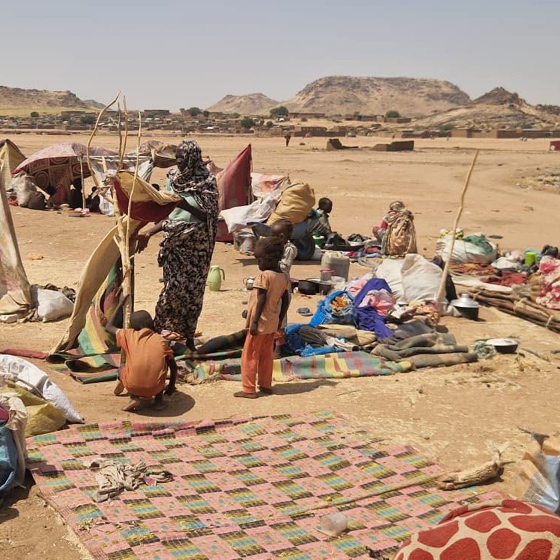 The image depicts a temporary settlement, likely a displaced persons camp or informal shelter in a rugged landscape. Several makeshift tents and tarps are set up, providing shelter for the residents. In the foreground, a woman is seen holding a structure, possibly a pole or support for one of the tents, while a child stands nearby. The ground is scattered with blankets, bags, and various belongings, suggesting a living area that is communal and somewhat crowded. The background shows a barren landscape with hills or rocky formations, indicating a harsh environment.