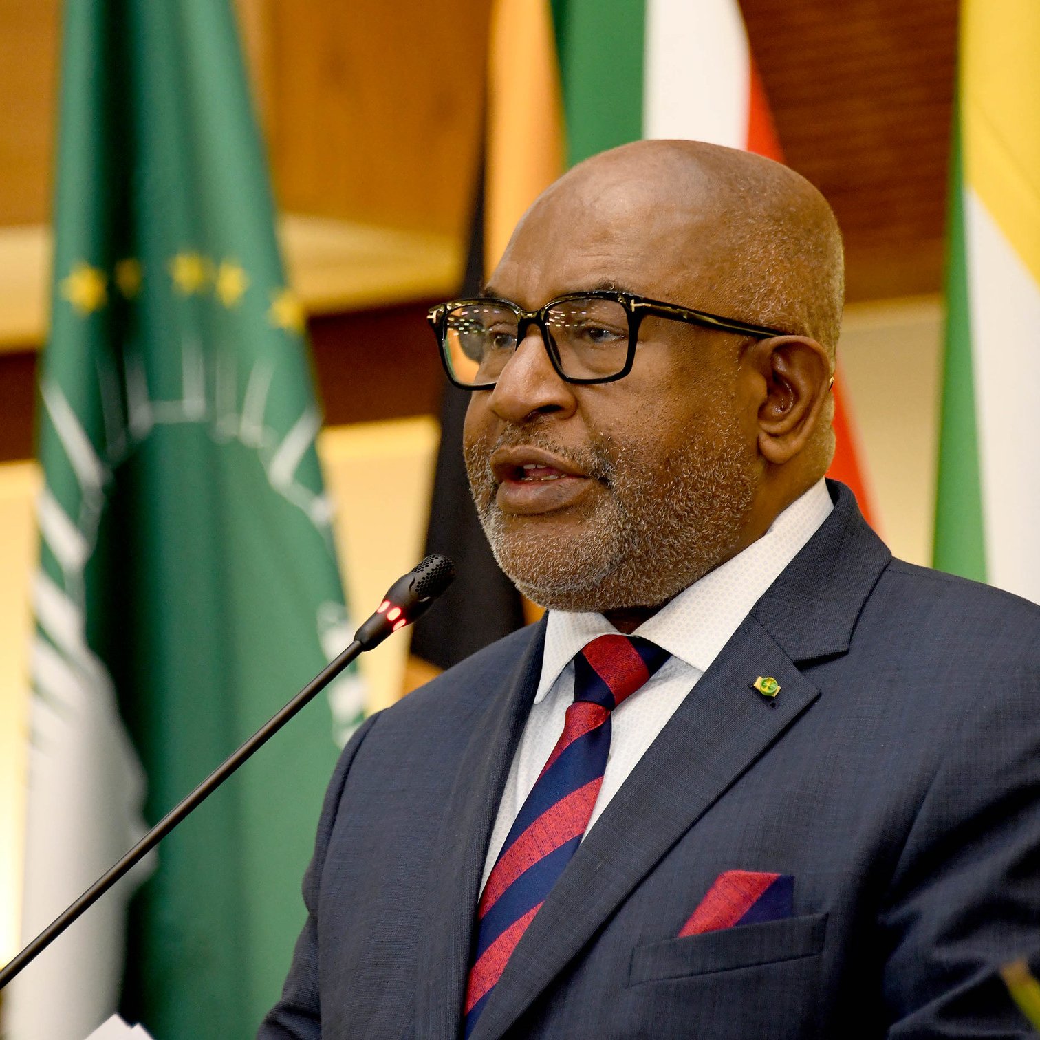 L'image représente un homme en train de prendre la parole lors d'un événement officiel. Il porte un costume sombre avec une chemise blanche et une cravate rayée, dans des tons de rouge et bleu. Ses lunettes sur le nez lui donnent une allure distinguée. Il se tient près d'un microphone, semblant s'adresser à un public. En arrière-plan, on peut apercevoir des drapeaux représentant différents pays, ce qui suggère que l'événement a une portée internationale. L'atmosphère semble solennelle et officielle, marquée par un éclairage chaleureux.