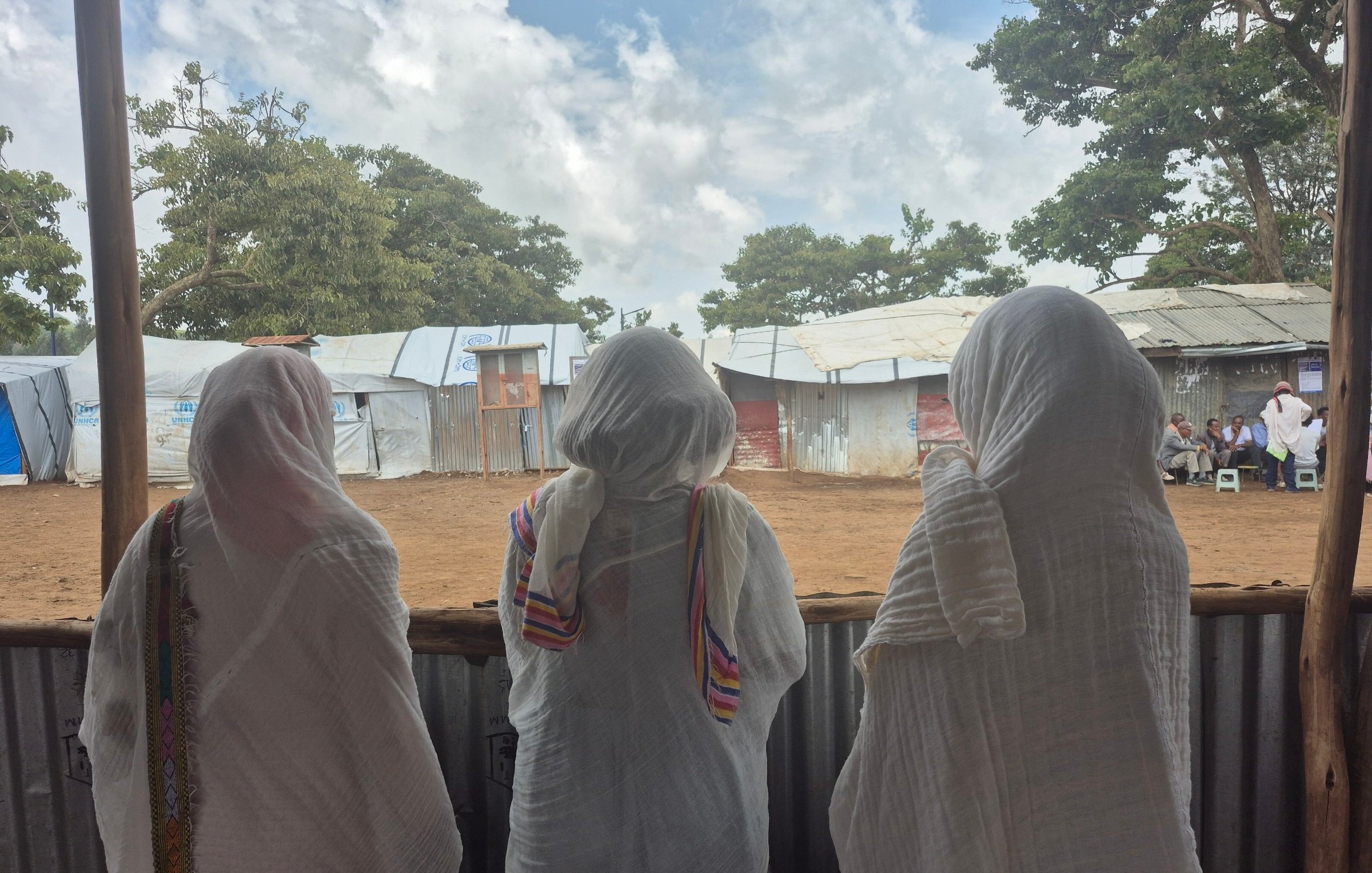 L'image montre trois femmes de dos, se tenant côte à côte, regardant à travers une structure en bois ouverte. Elles portent des vêtements longs et légers. Leurs têtes sont couvertes de foulards, ce qui suggère un contexte culturel ou religieux. Au loin, on aperçoit des abris temporaires, probablement des tentes ou des structures en tôles. Le ciel est partiellement nuageux, créant une atmosphère paisible. L'environnement évoque un lieu de rassemblement ou d'attente, avec un sol en terre battue. La scène semble calme, laissant entrevoir un moment de contemplation ou de partage entre ces femmes.