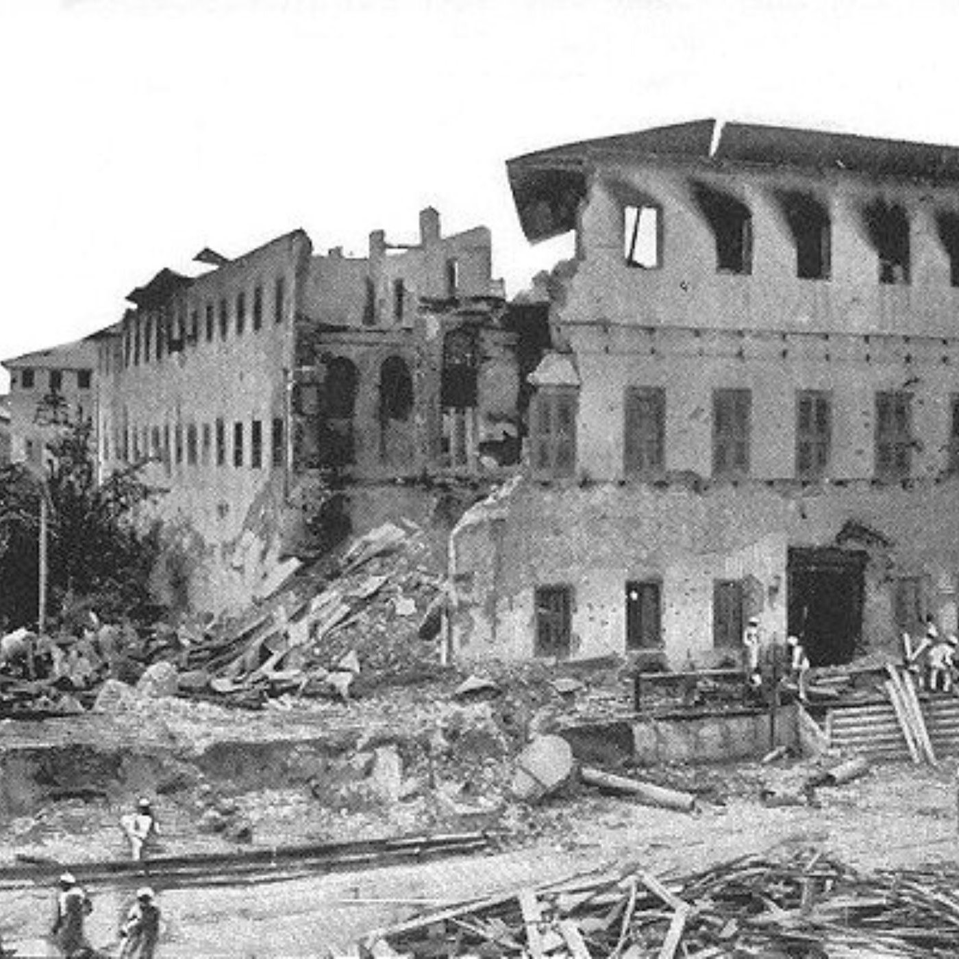 L'image montre un bâtiment sévèrement endommagé, probablement à la suite d'un tremblement de terre ou d'une catastrophe. La façade du bâtiment est partiellement effondrée, révélant des murs fissurés et des fenêtres brisées. Des débris sont éparpillés autour, créant un paysage chaotique. Au premier plan, on peut voir un groupe de personnes qui semblent examiner les dégâts, échangeant probablement leurs impressions. L'environnement est empreint d'une atmosphère de désolation, illustrant les conséquences d'un événement tragique sur l'architecture et la communauté environnante.