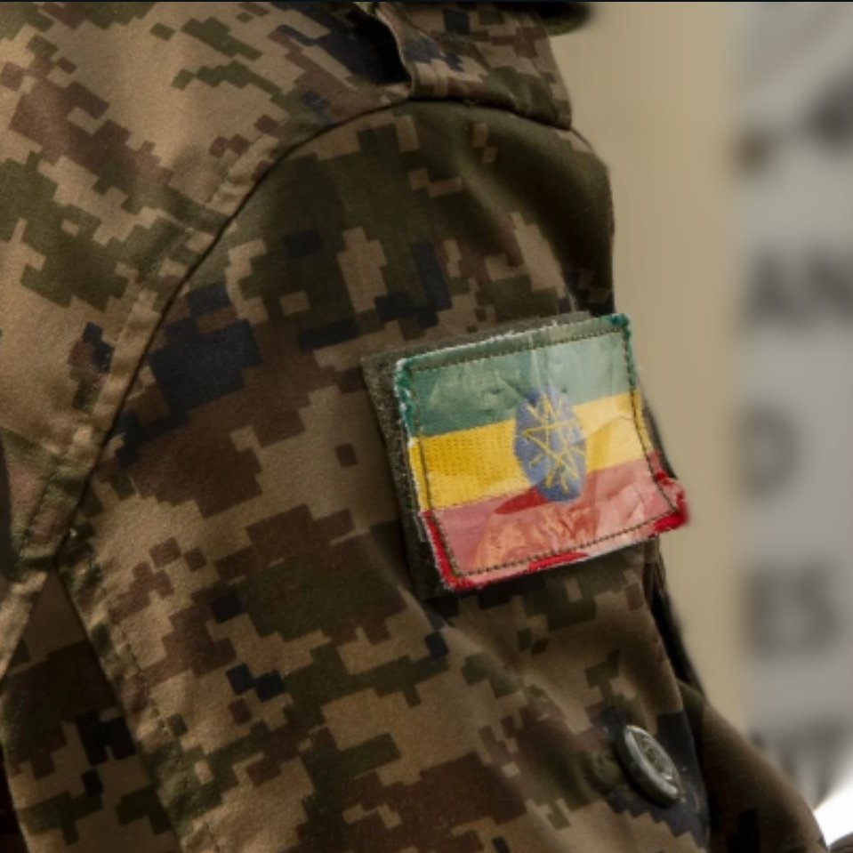The image features a close-up of a military uniform with a camouflage pattern. Prominently displayed on the arm is a patch showing the flag of Ethiopia, which consists of horizontal stripes in green, yellow, and red, with a blue circle and a yellow star in the center. The fabric of the patch appears worn, adding to its character.