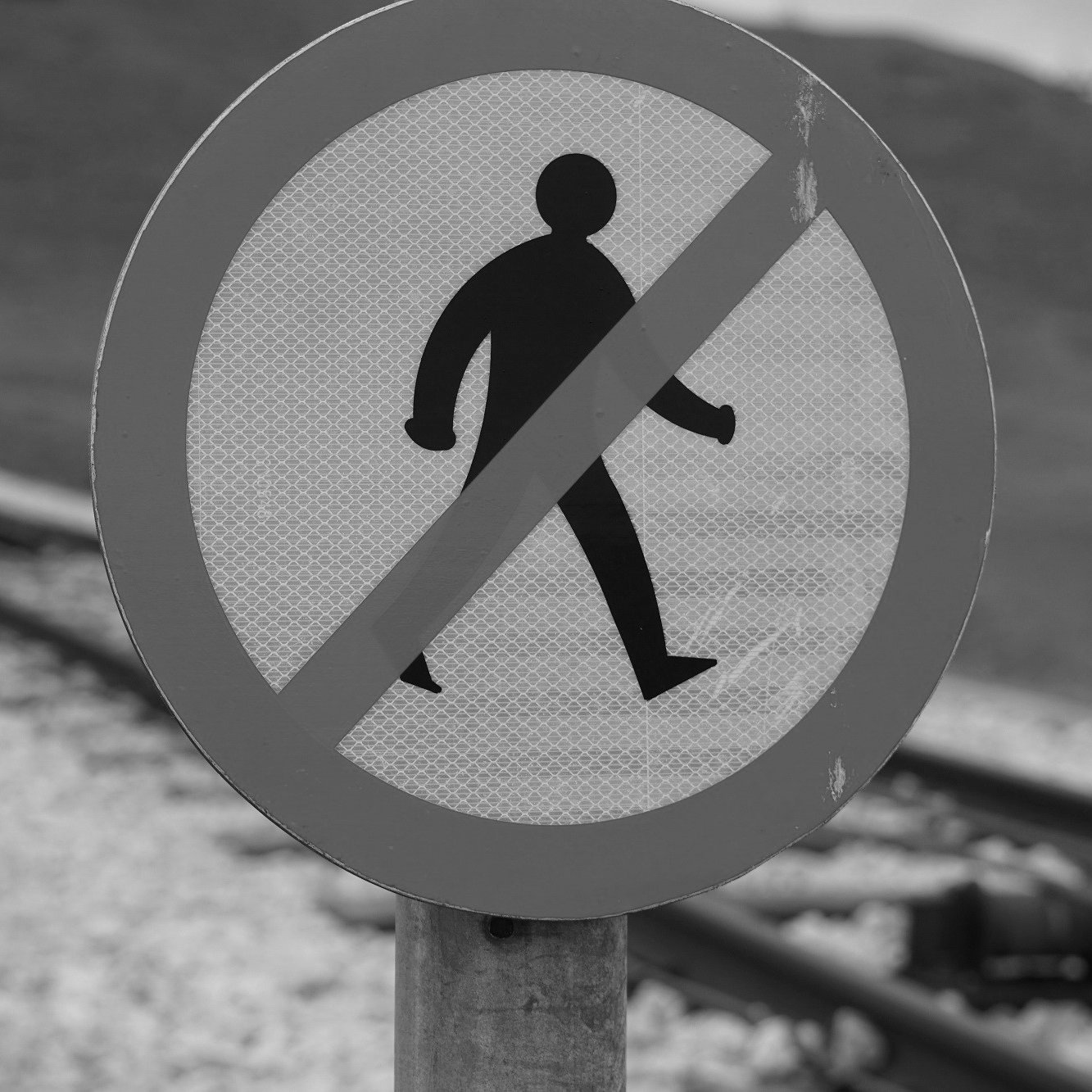 L'image présente un panneau de signalisation circulaire à fond blanc avec une silhouette noire d'une personne marchant. Une barre rouge diagonale traverse le panneau, indiquant qu'il est interdit de marcher ou d'accéder à cette zone. En arrière-plan, on devine des voies ferrées, suggérant que ce panneau est situé à proximité d'une zone ferroviaire, où l'accès est restreint pour des raisons de sécurité. L'image est en noir et blanc, ajoutant un contraste marqué.