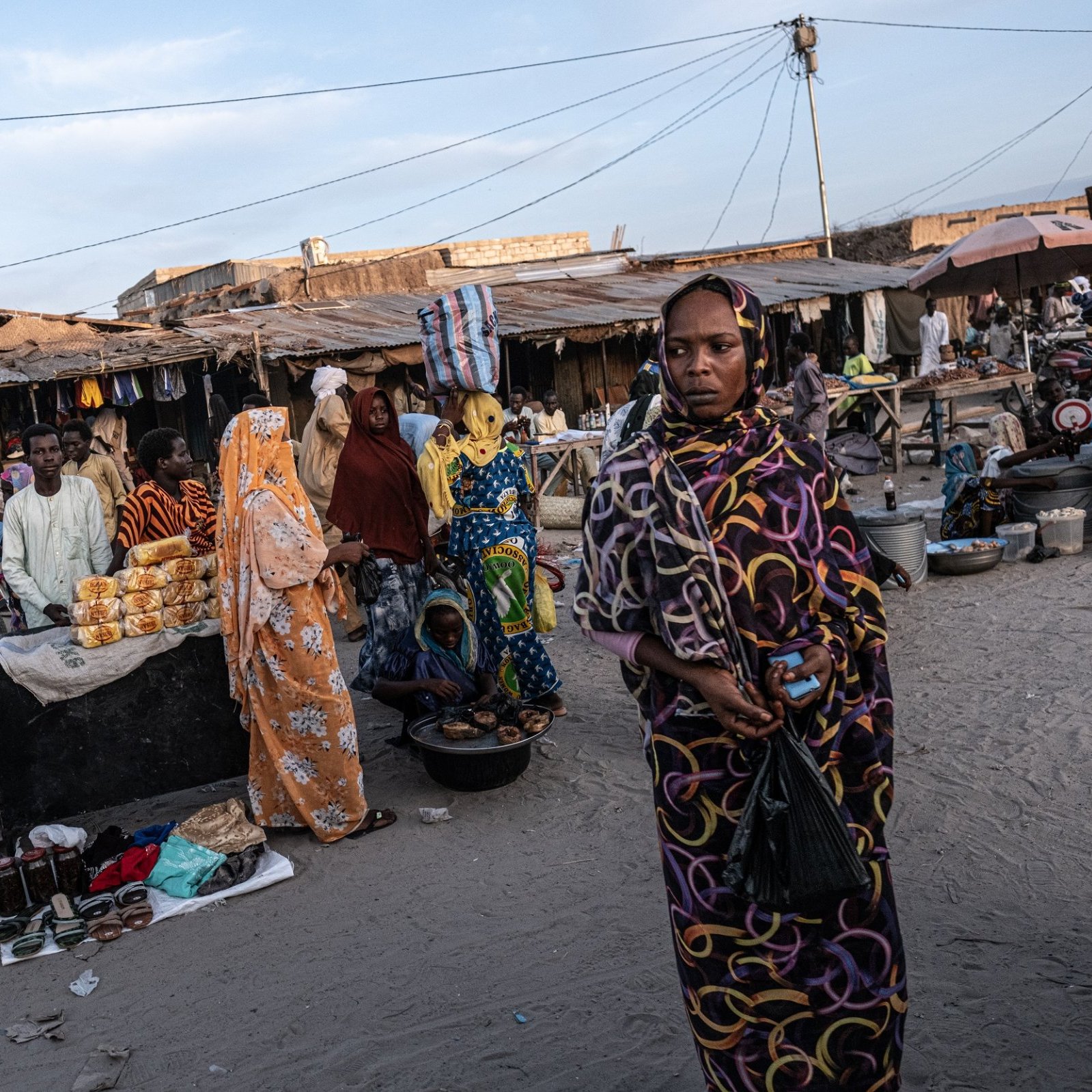 Dans cette scène animée d'un marché, on peut ressentir une atmosphère vibrante. Le sol est recouvert de sable, et plusieurs stands sont disposés tout autour. Des gens sont rassemblés, certains vêtus de vêtements colorés, offrant divers produits alimentaires, tels que des fruits et des pâtisseries. Les femmes, en particulier, attirent l'attention avec leurs robes chatoyantes et leurs voiles. On peut aussi entendre des murmures de conversations, des rires et le bruit de petits véhicules, comme des motos, qui se déplacent à travers le marché. L'air est imprégné d'odeurs variées de nourriture épicée et de douceur, créant une expérience sensorielle unique.