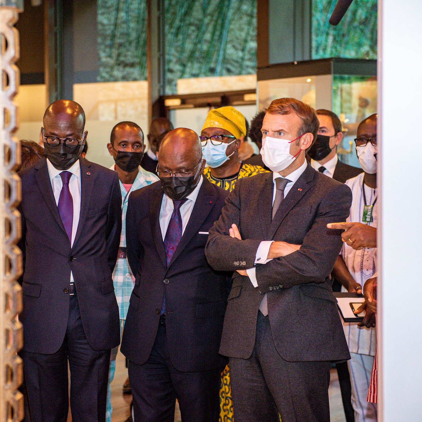 L'image montre un groupe de personnes, dont trois hommes, en train d'observer un objet d'art ou une exposition. Les hommes sont habillés de costumes formels, avec deux en costumes sombres et un en costume gris avec une cravate. Ils affichent des expressions de concentration et d'intérêt. À l'arrière-plan, on perçoit des éléments culturels, probablement des œuvres d'art ou des artifacts, créant une ambiance muséale. Les visiteurs portent tous des masques, ce qui indique qu'ils se trouvent dans un contexte où cela est nécessaire, peut-être en raison de préoccupations sanitaires. Le cadre est bien éclairé, mettant en valeur l'importance de l'exposition.