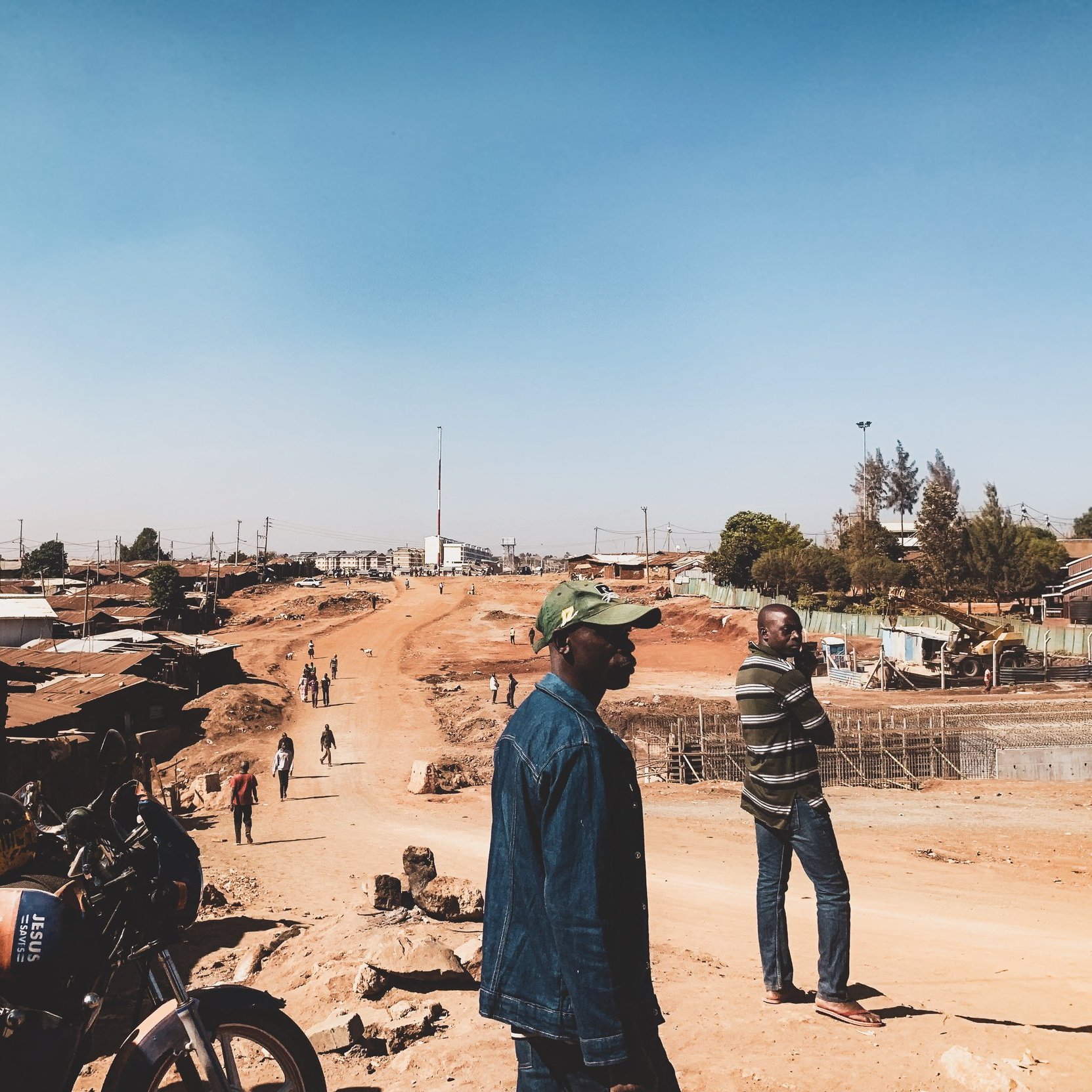 Dans cette image, nous voyons une scène d'une rue en construction dans une zone urbaine. Le ciel est dégagé et d'un bleu clair. Au premier plan, deux hommes se tiennent debout : l'un porte une veste en jean et une casquette, tandis que l'autre est vêtu d'un sweater rayé. À droite, on aperçoit un chemin de terre, entouré de bâtiments en dur et d'habitations simples. À gauche, une moto est garée, et plusieurs personnes se déplacent le long de la route. L'atmosphère semble calme, mais l'environnement indique une activité en cours avec des travaux de construction en arrière-plan.