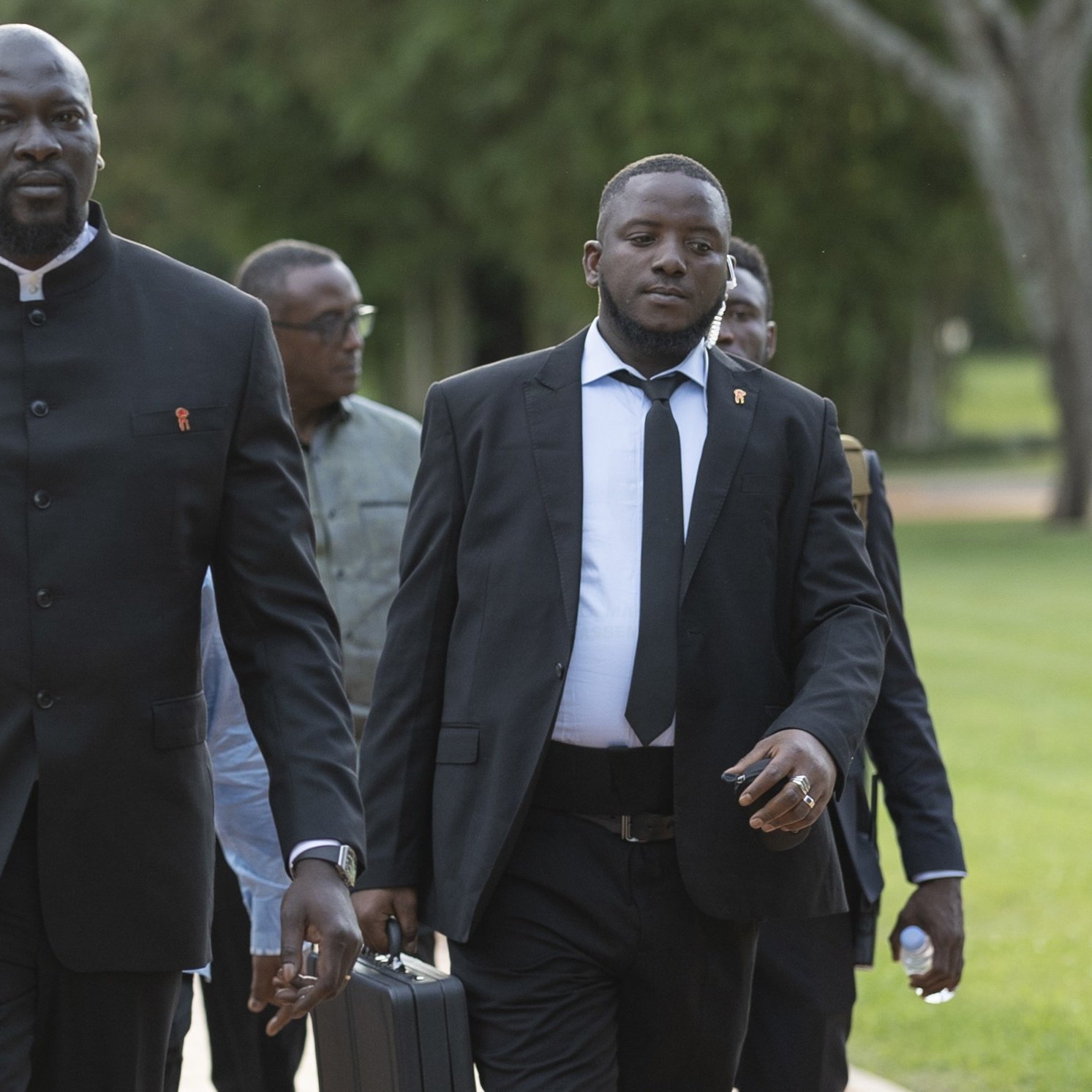 The image depicts a group of several men walking together in a park-like setting. The man in the foreground, dressed in a black suit with a high collar, appears confident and focused. He is accompanied by another man wearing a black suit and tie, who carries a briefcase. The background features lush greenery, suggesting an outdoor event or gathering. Other men can be seen following closely behind, contributing to a formal yet relaxed atmosphere.