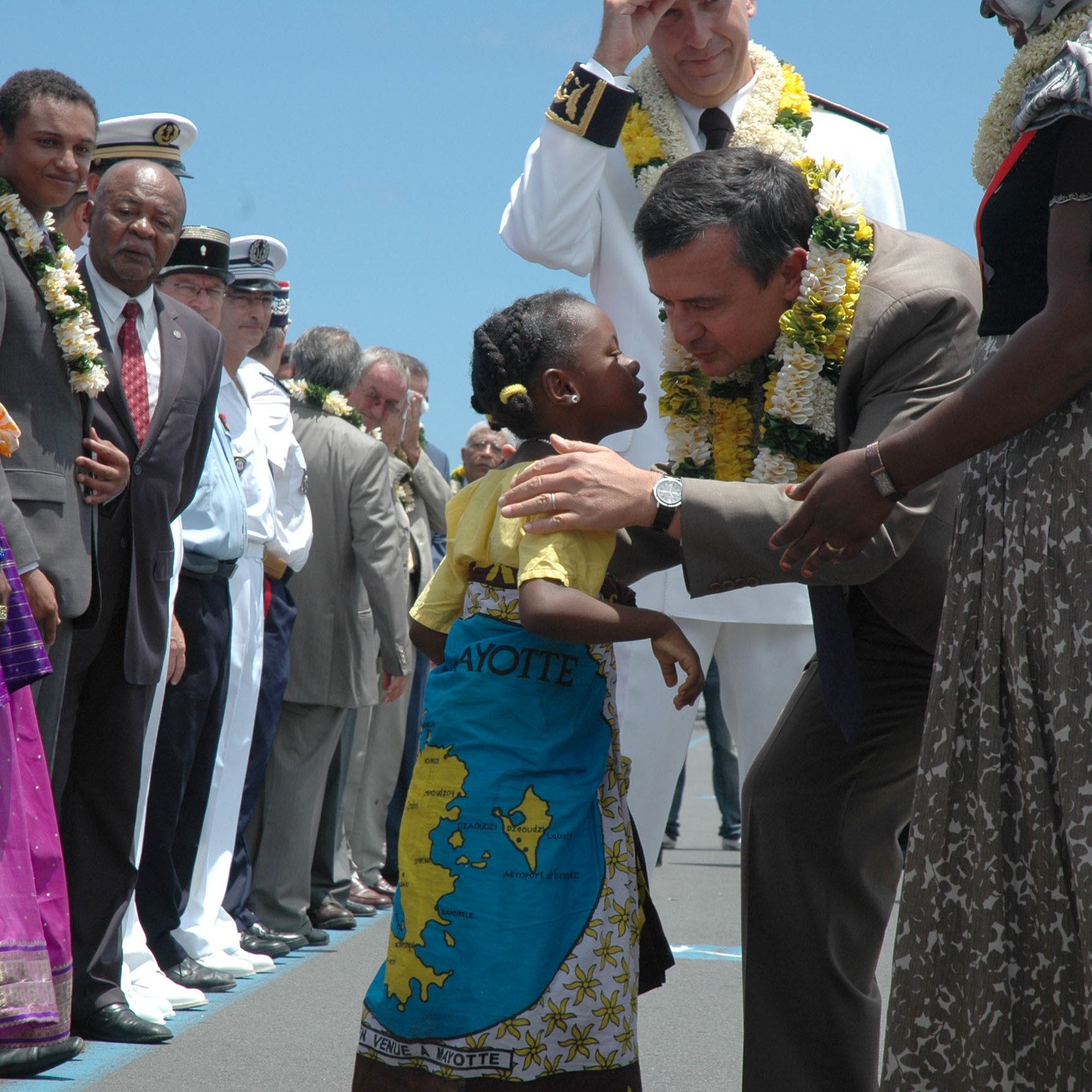 L'image représente une scène d'accueil vibrant sur un aéroport. Au premier plan, un homme en costard se penche pour embrasser une petite fille qui porte une robe colorée et un paréo. La fillette a un grand sourire et semble très joyeuse. Autour d'eux, plusieurs autres personnes, dont des adultes en vêtements traditionnels et militaires, les observent avec des expressions bienveillantes. Certains présents portent des guirlandes de fleurs autour du cou, ajoutant une touche festive à l'atmosphère. En arrière-plan, le ciel est clair, suggérant une journée ensoleillée. L'ensemble de la scène dégage une impression de chaleur et de convivialité.