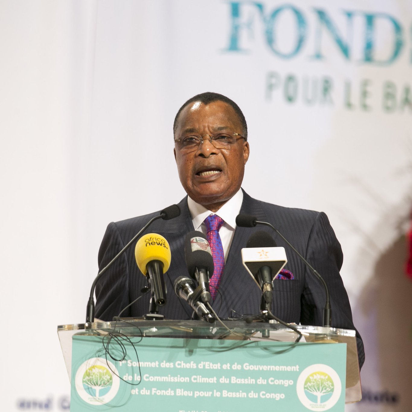 L'image montre un homme, probablement un leader ou un représentant politique, s'exprimant devant un public. Il porte un costume sombre avec une cravate rose et semble être en train de donner un discours. Devant lui, il y a un podium avec plusieurs microphones et un panneau indiquant "Fonds Bleu pour le Bassin du Congo". En arrière-plan, on peut voir des éléments décoratifs qui suggèrent un événement officiel, avec des logos et des affiches liés au thème de la conservation et du développement. L'atmosphère semble sérieuse, indiquant l'importance de l'événement.