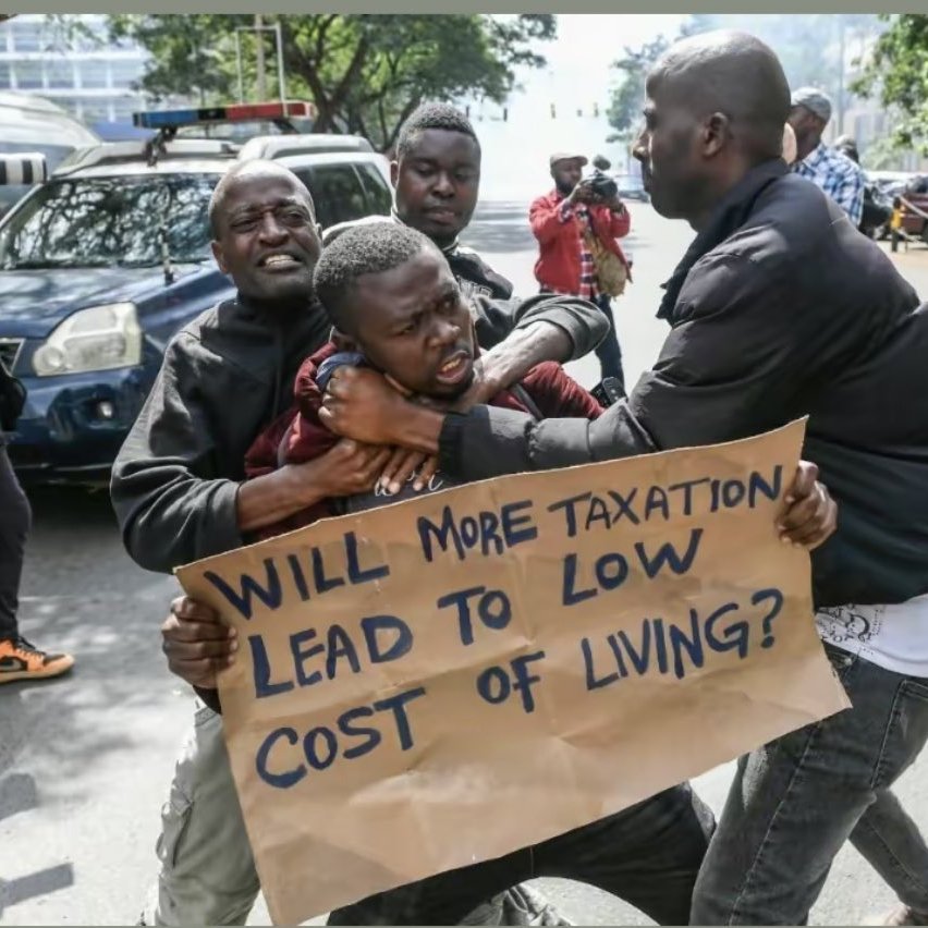 L'image montre une scène de protestation urbaine. Au premier plan, deux hommes sont engagés dans une altercation. L'un d'eux semble essayer de saisir l'autre qui tient une pancarte. Sur cette pancarte, on peut lire une question écrite en lettres noires : "WILL MORE TAXATION LEAD TO LOW COST OF LIVING?" (Plus de taxation conduira-t-elle à un coût de la vie plus bas ?). En arrière-plan, on aperçoit d'autres personnes, certaines les regardent, tandis que d'autres semblent prendre des photos. Des véhicules sont garés sur le côté, ajoutant à l'ambiance animée de la scène. Les expressions des hommes montrent des émotions vives, reflétant l'intensité de la situation.