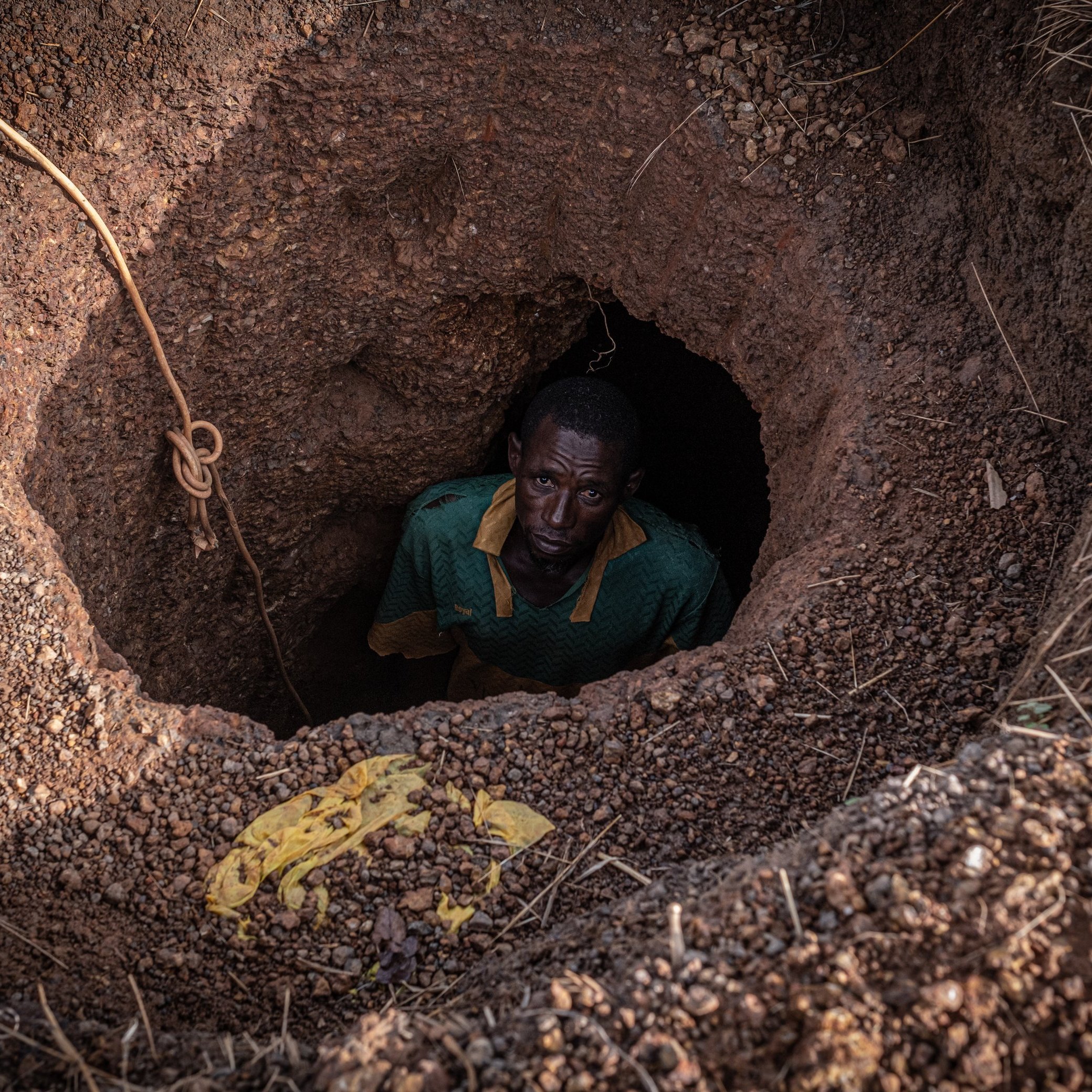 Dans cette image, nous voyons un homme qui se tient au fond d'un trou creusé dans le sol. Le trou a une forme ronde, et les parois sont en terre brune, légèrement humides et irrégulières. L'homme, vêtu d'un maillot vert avec un col jaune, regarde vers le haut avec une expression sérieuse. Son visage est en partie dans l'ombre, mais son regard est visible. Autour du trou, on peut apercevoir des gravats et des brindilles qui ajoutent un aspect naturel et brut à l'environnement. La scène évoque un sentiment d'isolement et de travail ardu.