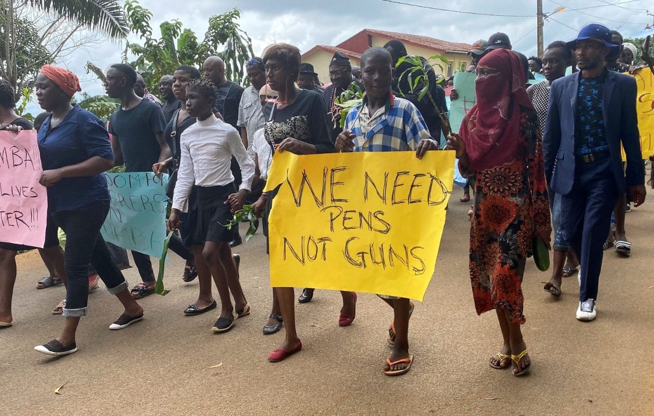 Dans cette image, un groupe de personnes marche ensemble dans une rue. On peut entendre des voix et des chants de solidarité. Les participants tiennent des pancartes aux couleurs vives. Certaines pancartes affichent des messages puissants comme "WE NEED PENS NOT GUNS", exprimant une demande pour la paix et l'éducation plutôt que pour la violence. Les manifestants sont vêtus de différents styles de vêtements, allant de tenues formelles à des vêtements décontractés. En arrière-plan, on aperçoit des maisons et des arbres qui ajoutent un cadre naturel à cette scène de protestation pacifique. L'atmosphère est déterminée et engagée, reflet d'un désir de changement et d'un message fort pour la communauté.
