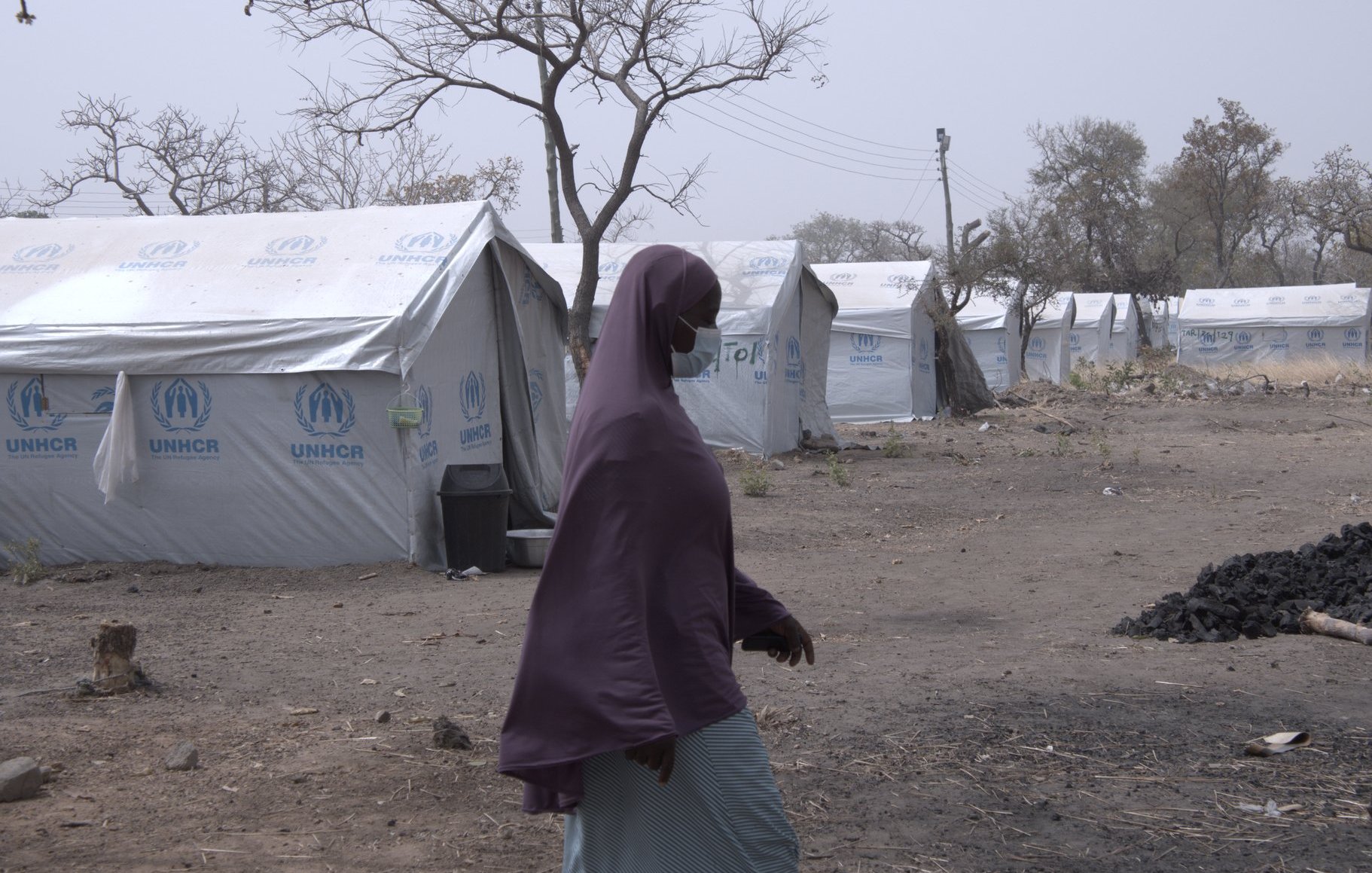 L'image montre un camp de réfugiés, caractérisé par plusieurs tentes en toile blanches et bleues, portant le logo de l'UNHCR. Au premier plan, une femme marche, vêtue d'un voile mauve et d'une robe claire, se dirigeant vers l'une des tentes. Le paysage environnant est aride et désolé, avec quelques arbres dénudés, suggérant un environnement difficile. En arrière-plan, des lignes électriques sont visibles, ce qui indique une certaine infrastructure. L'atmosphère semble calme, mais il y a une sensation de précarité et de résilience.