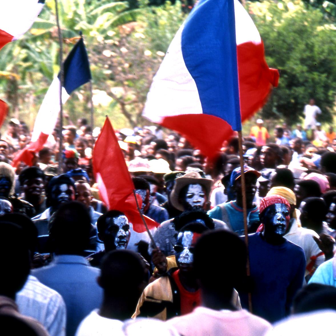 L'image représente une foule dense lors d'un événement, probablement un rassemblement ou une manifestation. Au premier plan, on peut voir des personnes brandissant des drapeaux français, avec des couleurs bleu, blanc et rouge. Certains membres de la foule portent des maquillages colorés sur le visage, ajoutant une dimension festive ou militante à la scène. Les visages sont à la fois heureux et engagés, témoignant d'une ambiance dynamique. En arrière-plan, des arbres et de la verdure suggèrent que l'événement se déroule dans un espace extérieur, peut-être dans un endroit tropical ou rural. L'ensemble dégage une forte impression de solidarité et de passion collective.