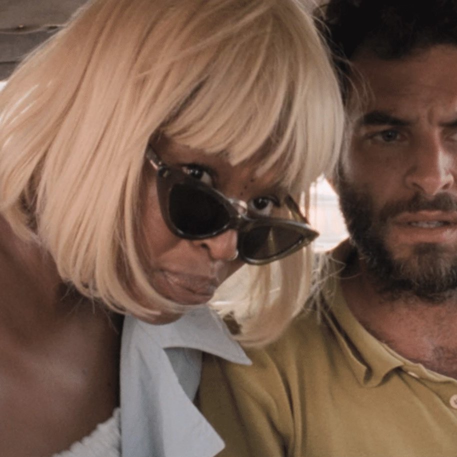 Dans cette image, on voit deux personnes assises à l'arrière d'un véhicule. À gauche, une femme d'une trentaine d'années a les cheveux blonds et courts, coiffés en une coupe élégante. Elle porte de grandes lunettes de soleil noires et une tenue légère. Son expression est attentive, presque inquisitrice. À droite, un homme d'une trentaine d'années également, a une barbe foncée et des cheveux bouclés. Il est vêtu d'une chemise de couleur terre. Son regard est préoccupé et intense, comme s'il était en train de réfléchir à quelque chose d'important. L'atmosphère dans l'image semble suggérer une conversation sérieuse ou un moment de tension partagée. Les sièges du véhicule sont en cuir ou en tissu sombre, et on peut apercevoir des objets non identifiables, peut-être des sacs ou des vêtements, à leurs pieds.