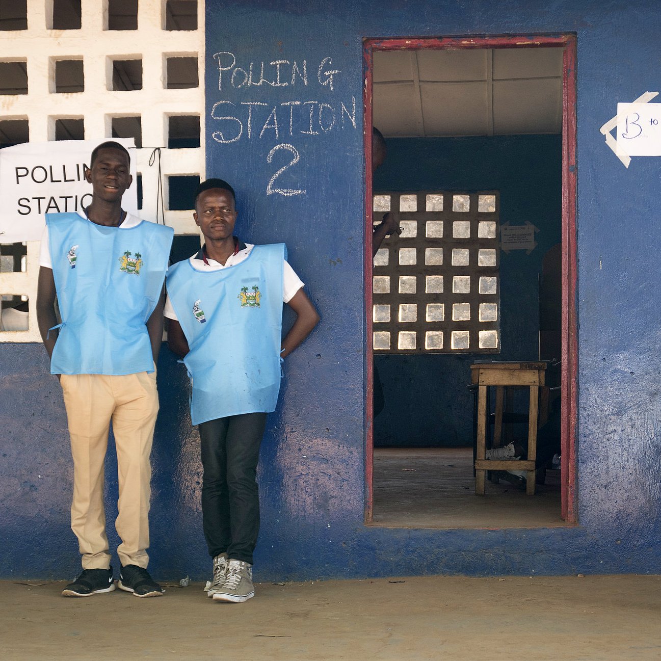 L'image montre une scène à l'extérieur d'un bureau de vote. À gauche, deux hommes portent des gilets bleus, probablement identifiant leur fonction d'observateurs ou de travailleurs électoraux. Ils se tiennent debout près de l'entrée, affichant une attitude accueillante. À droite, un autre homme est assis sur une chaise, paraissant détendu. L'environnement est coloré, avec des murs peints en bleu et des fenêtres comportant des grilles. Au-dessus de l'entrée, on peut voir le mot "Polling Station" écrit en anglais, indiquant qu'il s'agit d'un lieu de vote. Le sol est de terre battue, et l'atmosphère semble à la fois tranquille et sérieuse, typique d'un jour d'élection.