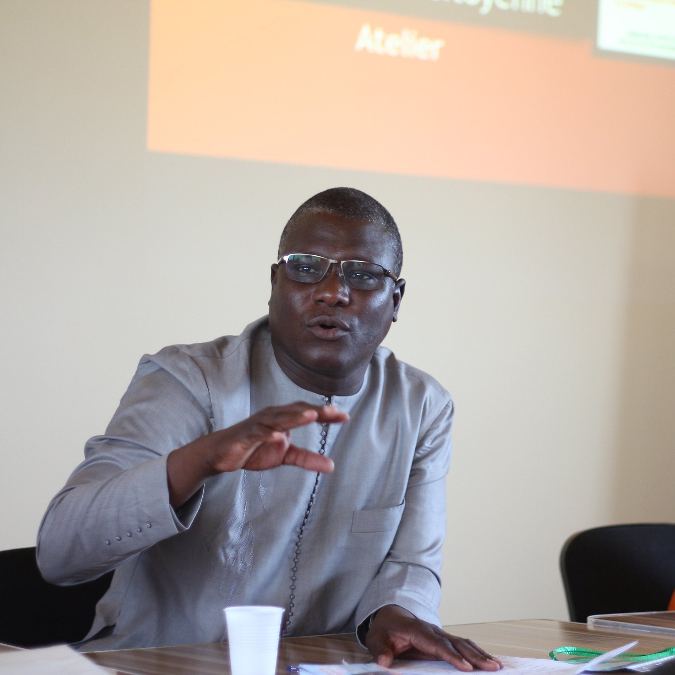 L'image montre un atelier où deux personnes sont assises à une table. À gauche, un homme, portant des lunettes et une tenue traditionnelle grise, gesticule avec une main tout en parlant. Son expression est engageante et dynamique, suggérant qu'il s'adresse à un groupe. À droite, une femme, habillée d'un t-shirt orange, écoute attentivement. Son visage montre une attitude attentive et intéressée. En arrière-plan, un écran est visible avec des textes, mais il est flou. L'atmosphère semble interactive et collaborative, indiquant un échange d'idées lors de l'atelier.