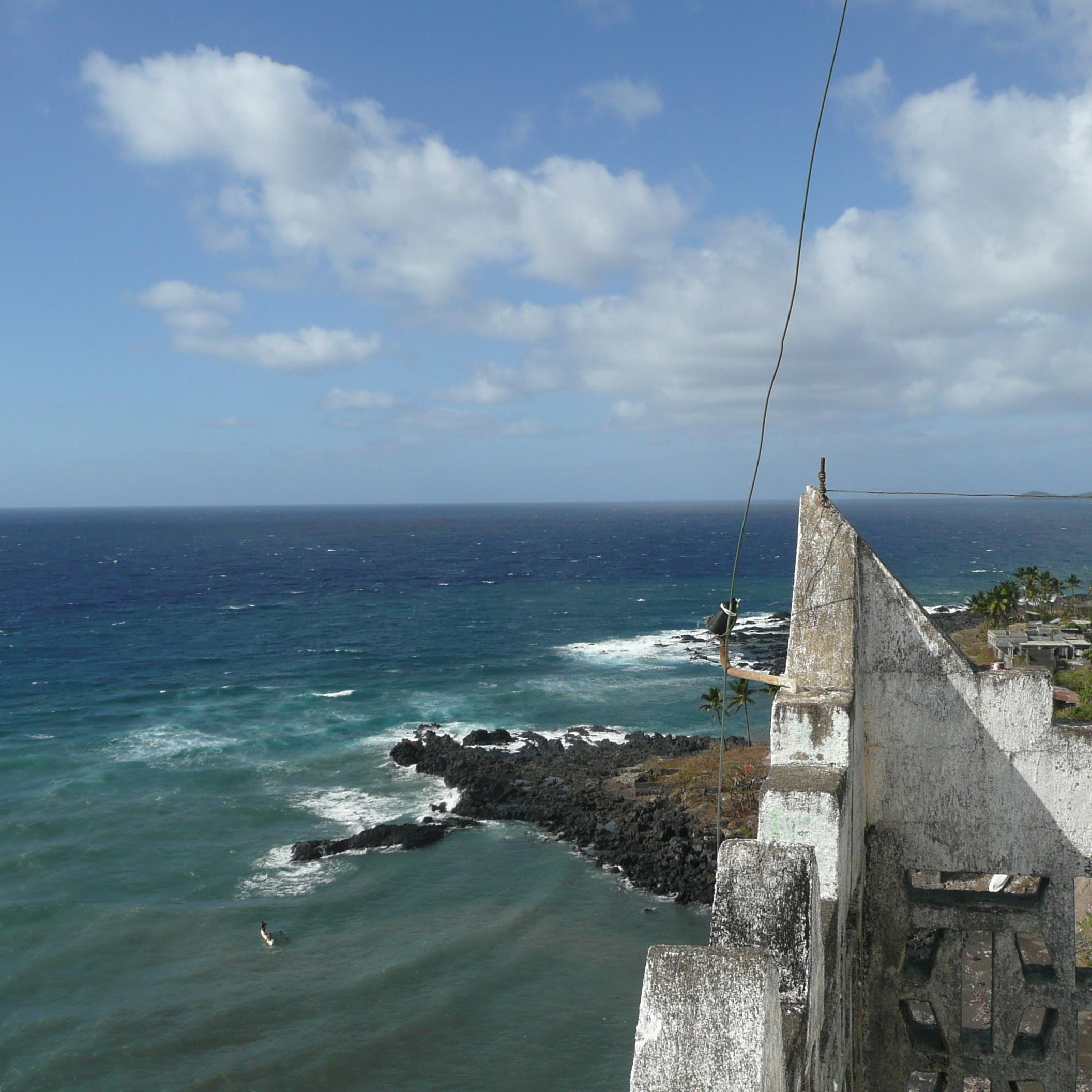 Imaginez-vous au sommet d'une vieille tour, où l'air est frais et légèrement salé. Devant vous s'étend un vaste océan d'un bleu profond, parsemé de vagues qui dansent et s'écrasent sur des rochers sombres. Au loin, vous pouvez percevoir la côtes, avec des palmiers se balançant doucement au gré du vent. Le ciel est parsemé de nuages blancs qui flottent, créant un jeu de lumière sur l'eau. Sur votre droite, des paysages verdoyants de collines se dessinent, ajoutant à la beauté sauvage de cet endroit. C'est un panorama paisible et apaisant, où la nature semble en harmonie avec l'horizon.