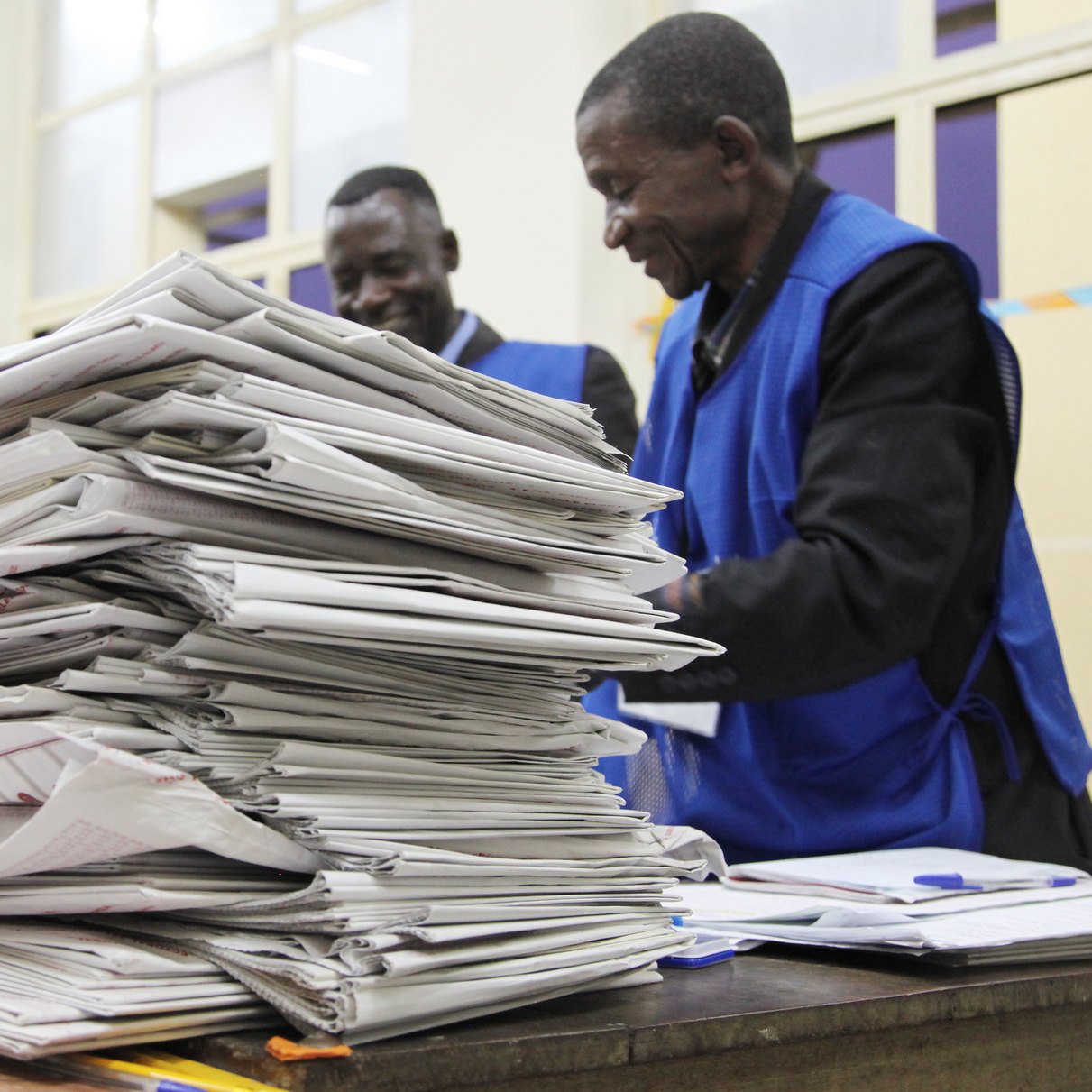 L'image montre une scène à l'intérieur d'un bureau de vote. Au premier plan, on aperçoit une grande pile de documents, probablement des bulletins de vote ou des formulaires électoraux, soigneusement empilés sur une table en bois. À l'arrière-plan, trois personnes en gilets bleus se trouvent près de la table, semblant discuter ou examiner les papiers. L'atmosphère est concentrée, suggérant une activité importante liée à un scrutin. La lumière éclaire la pièce, créant une ambiance sérieuse et professionnelle.