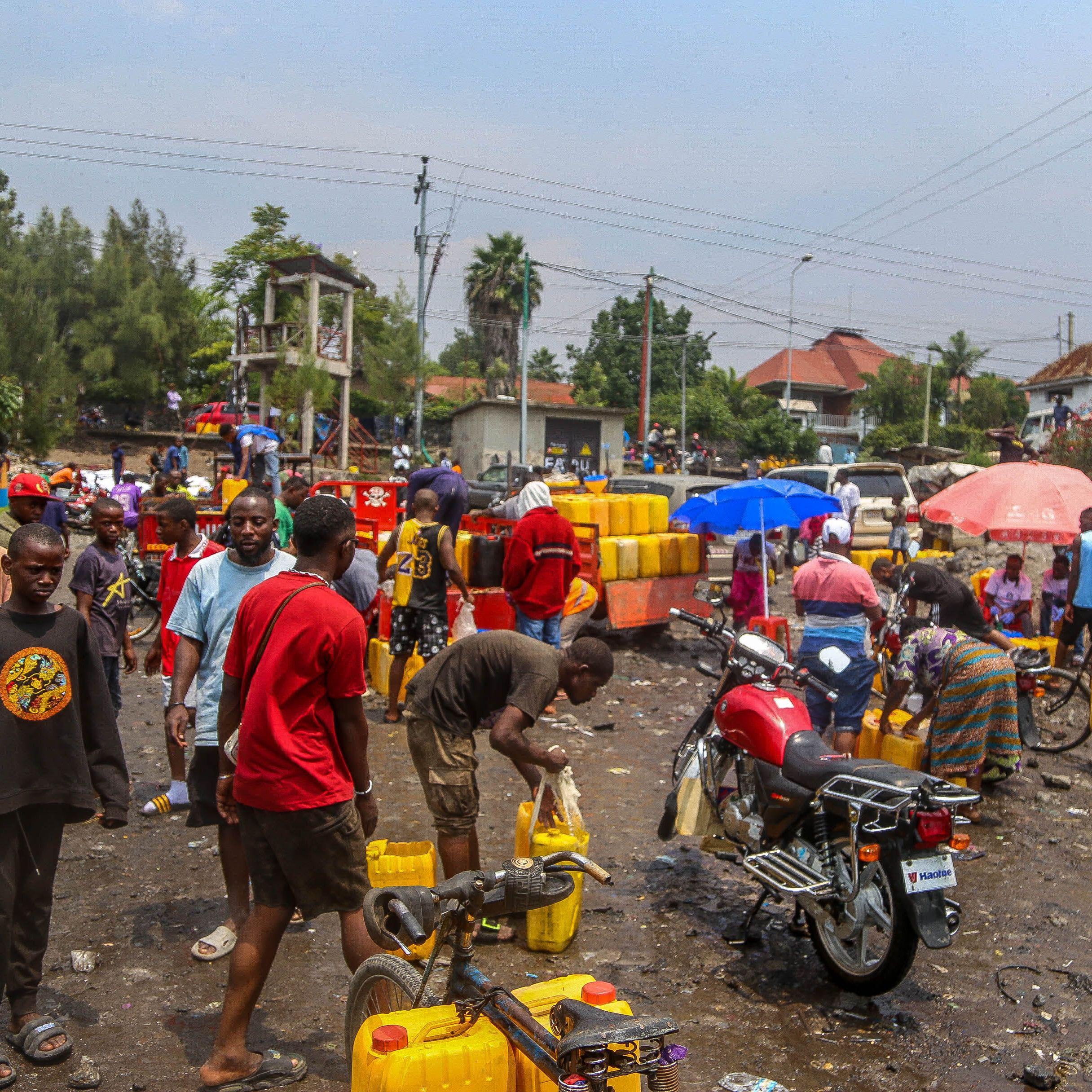 L'image représente une scène animée dans un quartier urbain. On peut imaginer un marché ou un point de distribution d'eau, où de nombreuses personnes sont rassemblées. Des hommes et des femmes, de divers âges, s'affairent autour de grands récipients jaunes qu'ils remplissent d'eau. Le sol semble en désordre, avec des déchets visibles, ajoutant une impression de négligence dans l'environnement. Des parapluies colorés sont déployés, fournissant un peu d'ombre sous un ciel légèrement nuageux. Au fond, on peut apercevoir des bâtiments et des arbres, tandis qu'un motard fait la queue avec sa moto, indiquant une activité routière environnante. L'atmosphère est dynamique, avec une forte interaction entre les gens.