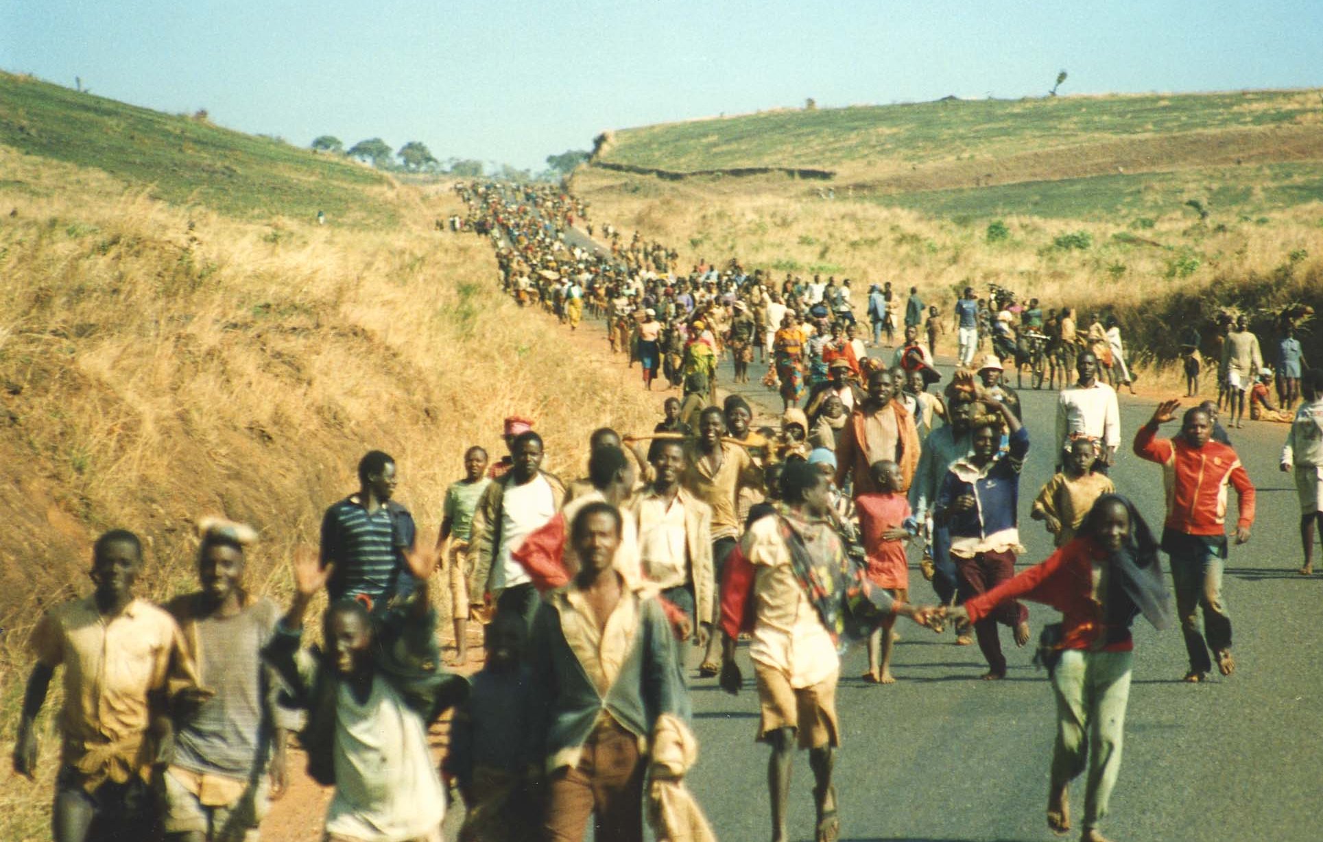 L'image montre une vaste foule de personnes marchant le long d'une route au milieu d'un paysage de collines sèches et herbeuses. Les individus, de différents âges et costumes, avancent ensemble, certains se tenant par la main ou portant des charges. L'atmosphère semble à la fois déterminée et solennelle, peut-être symbolisant un déplacement ou une migration collective. En arrière-plan, on aperçoit une étendue de terre vallonnée qui s'étend à perte de vue, ajoutant une dimension de profondeur à la scène. Le ciel est dégagé, laissant entrevoir un jour ensoleillé.