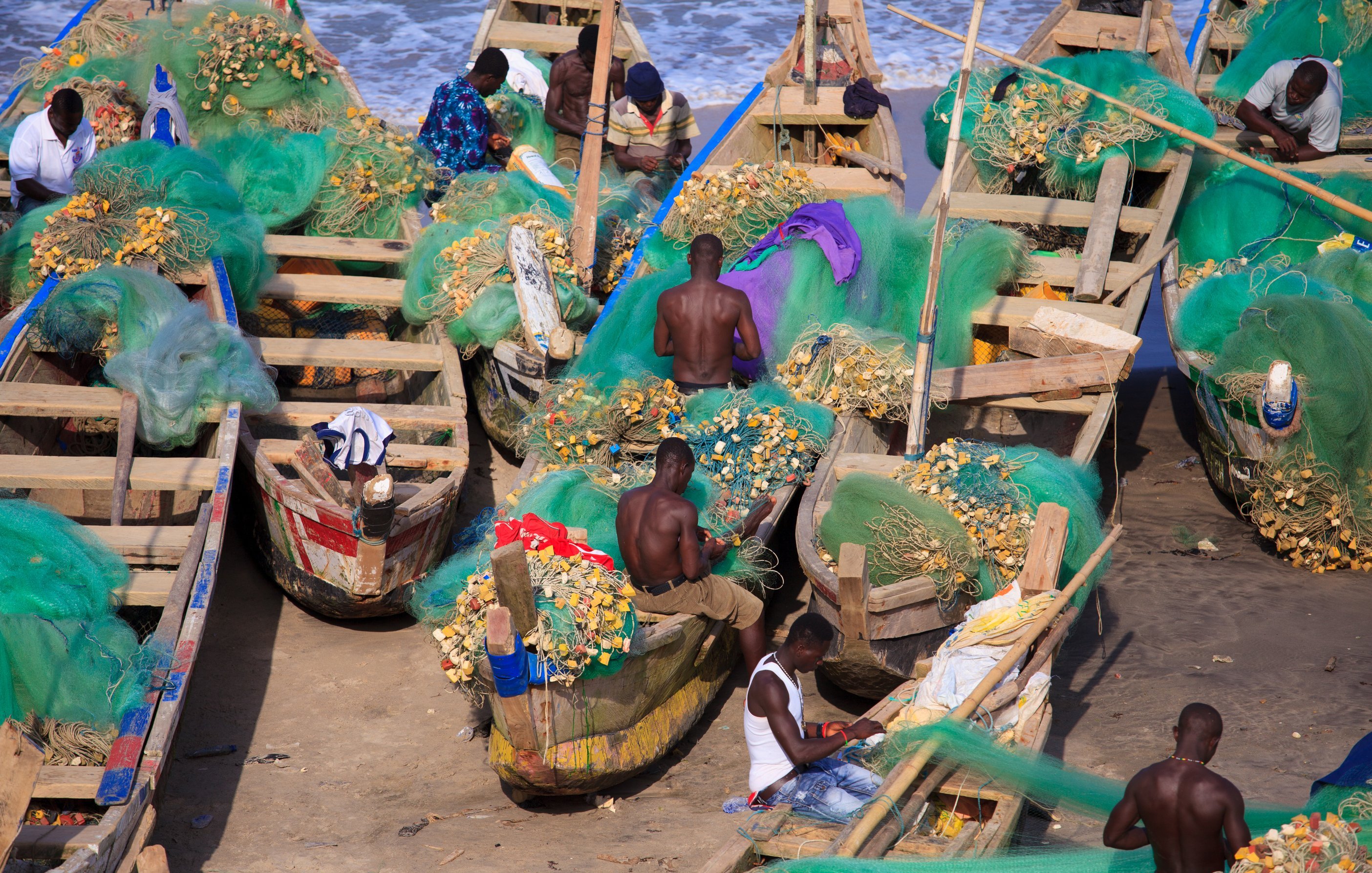 Cette image montre une scène vibrante en bord de mer, où plusieurs pêcheurs sont occupés à travailler sur des bateaux en bois. Les bateaux, peints de différentes couleurs, sont remplis de filets de pêche verts, ainsi que de coquillages et poisson. Les pêcheurs, principalement des hommes, sont torse nu et concentrés sur leurs tâches. L'arrière-plan révèle une mer calme avec des vagues douces, ajoutant une atmosphère de tranquillité à l'activité du port. L'air est imbibé de l'odeur de la mer et du bruit des vagues. Le soleil éclaire la scène, offrant une lumière chaude qui illumine le travail acharné des pêcheurs.