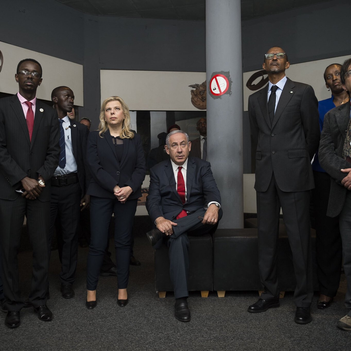L'image montre un groupe de personnes rassemblées dans une salle. Au centre, un homme est assis sur un canapé, vêtu d'un costume, tandis que les autres se tiennent debout autour de lui. Ils portent tous des costumes et ont des expressions sérieuses, suggérant une atmosphère formelle. L'environnement semble être une exposition ou un espace culturel, avec des œuvres d'art ou des objets exposés sur les murs. L'éclairage est doux, créant une ambiance réfléchie et attentive.