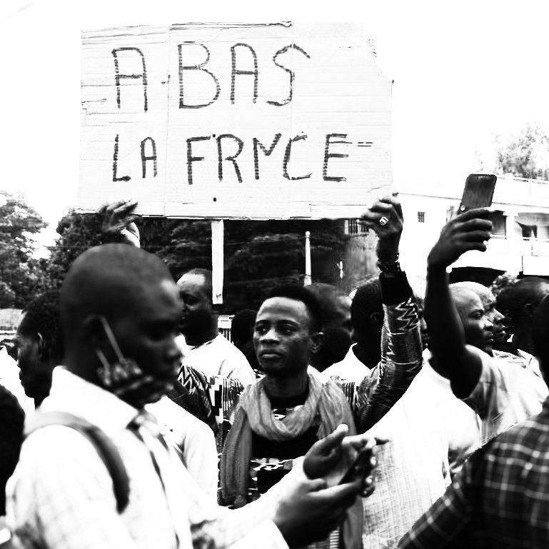 L'image montre une scène de manifestation. Au centre, une personne se tient debout, brandissant une grande pancarte sur laquelle est écrit "À BAS LA FRANCE". Cette phrase semble exprimer un sentiment de mécontentement. Autour de cette personne, un groupe d'hommes et de femmes montre diverses expressions, allant de la détermination à la colère. Certains tiennent des téléphones portables, prenant des photos ou des vidéos de la manifestation. L'arrière-plan est flou, mais on peut distinguer des bâtiments et des arbres. L'atmosphère générale dégage une forte émotion et un engagement collectif sur un sujet qui préoccupe les manifestants.