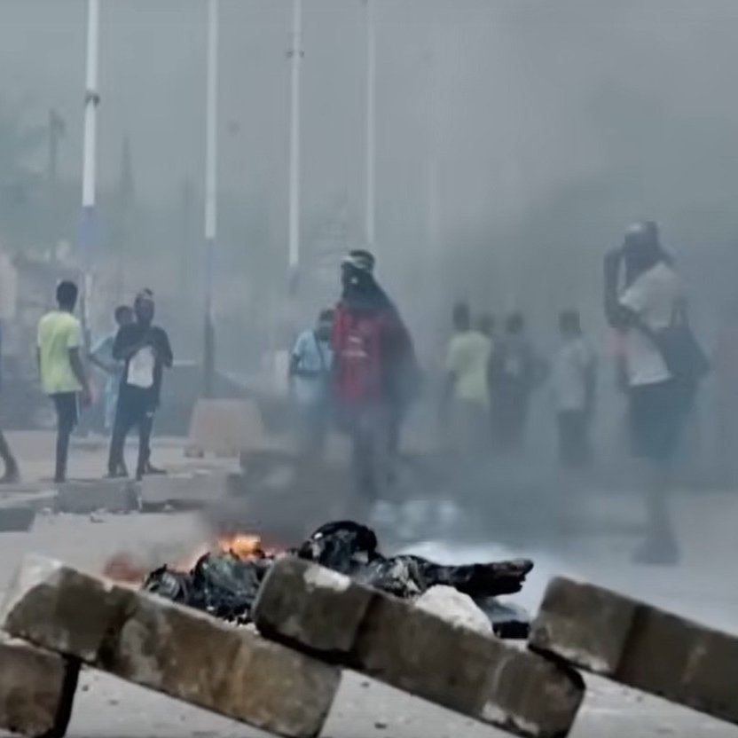 L'image montre une scène urbaine troublée. Au premier plan, plusieurs personnes courent rapidement, manifestant une impression d'urgence et de panique. En arrière-plan, de grands nuages de fumée noire s'élèvent dans l'air, indiquant un incendie. Des flammes orange et rouges jaillissent de barricades, suggérant une situation de conflit ou de désordre. Le décor urbain autour d'eux est flou, mais on peut discerner des bâtiments et des routes. L'atmosphère est tendue, évoquant un sentiment de danger et d'agitation.