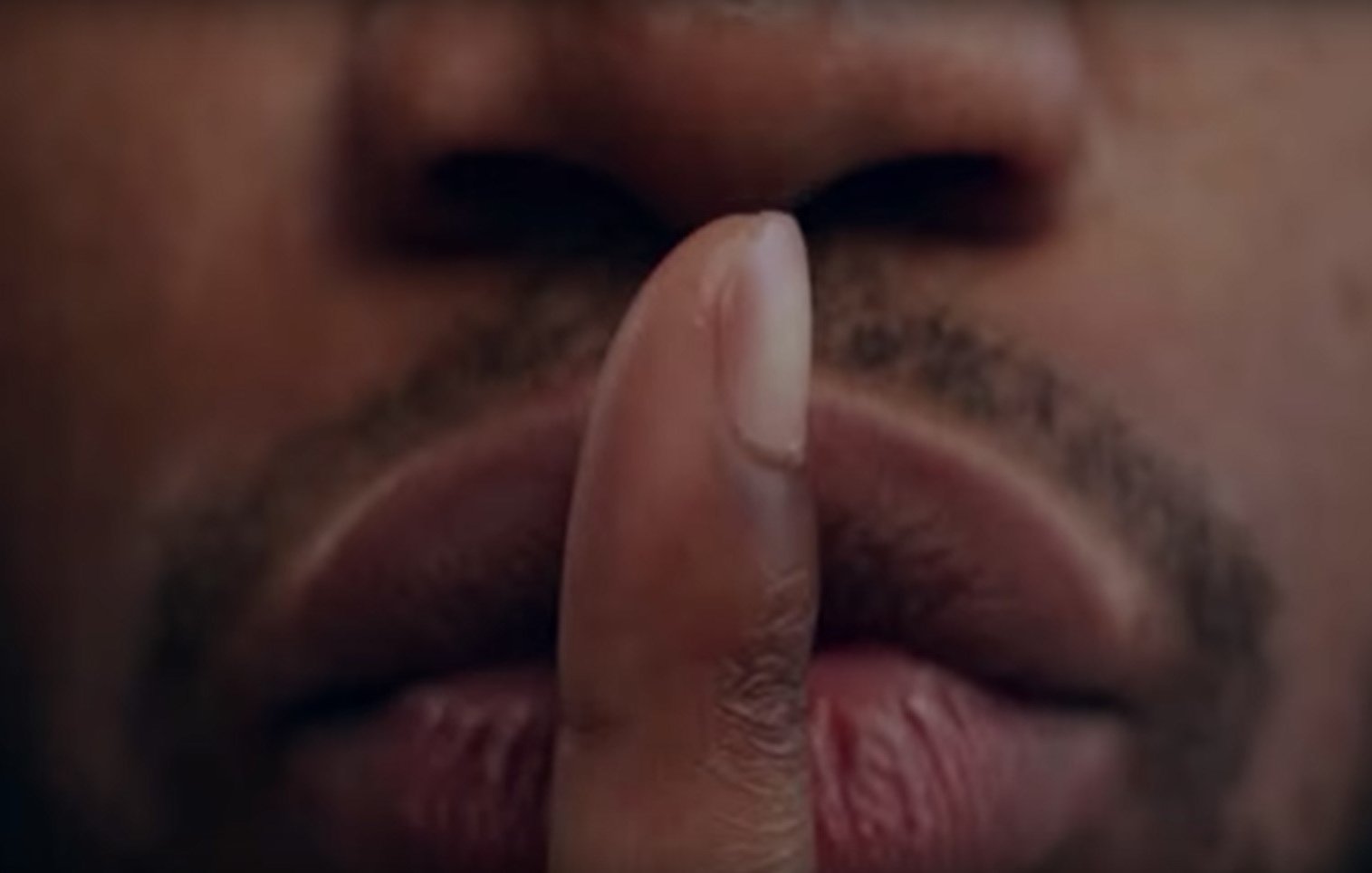 L'image présente un gros plan sur un visage, principalement la zone des lèvres et du nez. Un doigt est placé devant la bouche, formant le geste universel du « chut ». La peau est de teinte foncée, et les lèvres sont bien définies, affichant une légère brillance. Le regard de la personne est probablement dirigé vers l'extérieur de l'image, transmettant une impression de mystère ou de secret. L'arrière-plan est flou, ce qui met l'accent sur le geste et l'expression du visage.