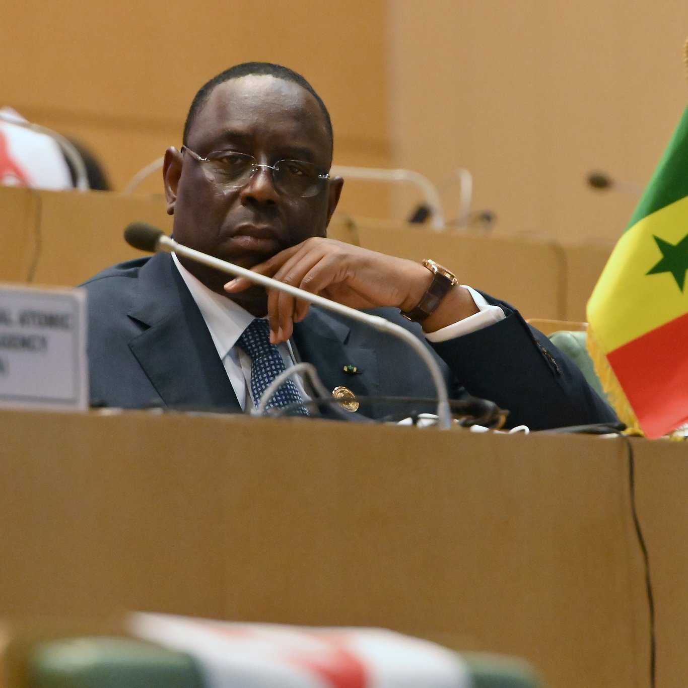 L'image montre un homme assis dans un cadre officiel, probablement lors d'une réunion ou d'une conférence. Il porte un costume sombre avec une chemise et une cravate, et affiche une expression pensive ou attentive. À sa droite, un petit drapeau du Sénégal est visible, avec les couleurs rouge, jaune et vert, ainsi que l'étoile verte au centre. On peut également apercevoir une bouteille d'eau à gauche et un badge ou un panneau indiquant "SENEGAL" devant lui. L'ambiance générale est sérieuse et concentrée, typique de discussions diplomatiques ou politiques.