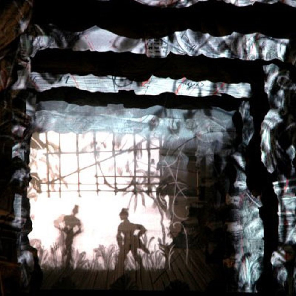 Imaginez une scène immersive où l'obscurité cède lentement la place à des formes délicates et intrigantes. Vous vous trouvez dans un espace structuré, semblable à un tunnel, dont les murs sont texturés, évoquant un rocher ou un vieux bois. Sur ces murs, des projections lumineuses créent des ombres dansantes, représentant des silhouettes de personnages en mouvement. Ces figures semblent interagir avec leur environnement, donnant vie à un récit visuel. Derrière ces silhouettes, on perçoit des éléments naturels, tels que des plantes ou des herbes, ajoutant une dimension organique à l'ensemble. L'atmosphère est à la fois mystérieuse et poétique, invitant à explorer les détails auditifs et tactiles qui pourraient compléter cette expérience. Vous pouvez presque sentir l'air ou l'humidité de l'espace, comme si vous étiez immergé dans un monde chargé de sensations et d'émotions.