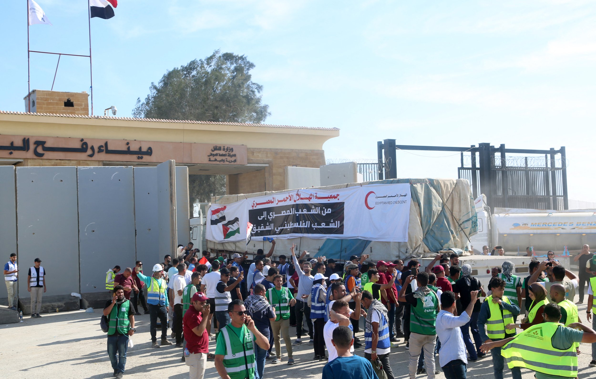 L'image montre une scène animée devant un point de passage, probablement dans une région à fort trafic. On aperçoit des personnes regroupées, certaines portant des gilets de sécurité ou des uniformes, tandis que d'autres tiennent des pancartes. En arrière-plan, on distingue un bâtiment gouvernemental avec des drapeaux en hauteur, ce qui suggère une ambiance officielle ou politique. Les gens semblent engagés dans une sorte de manifestation ou de rassemblement, discutant entre eux et brandissant des affiches qui expriment des revendications. L'atmosphère est festive mais sérieuse, marquée par le dynamisme des participants et les messages affichés.