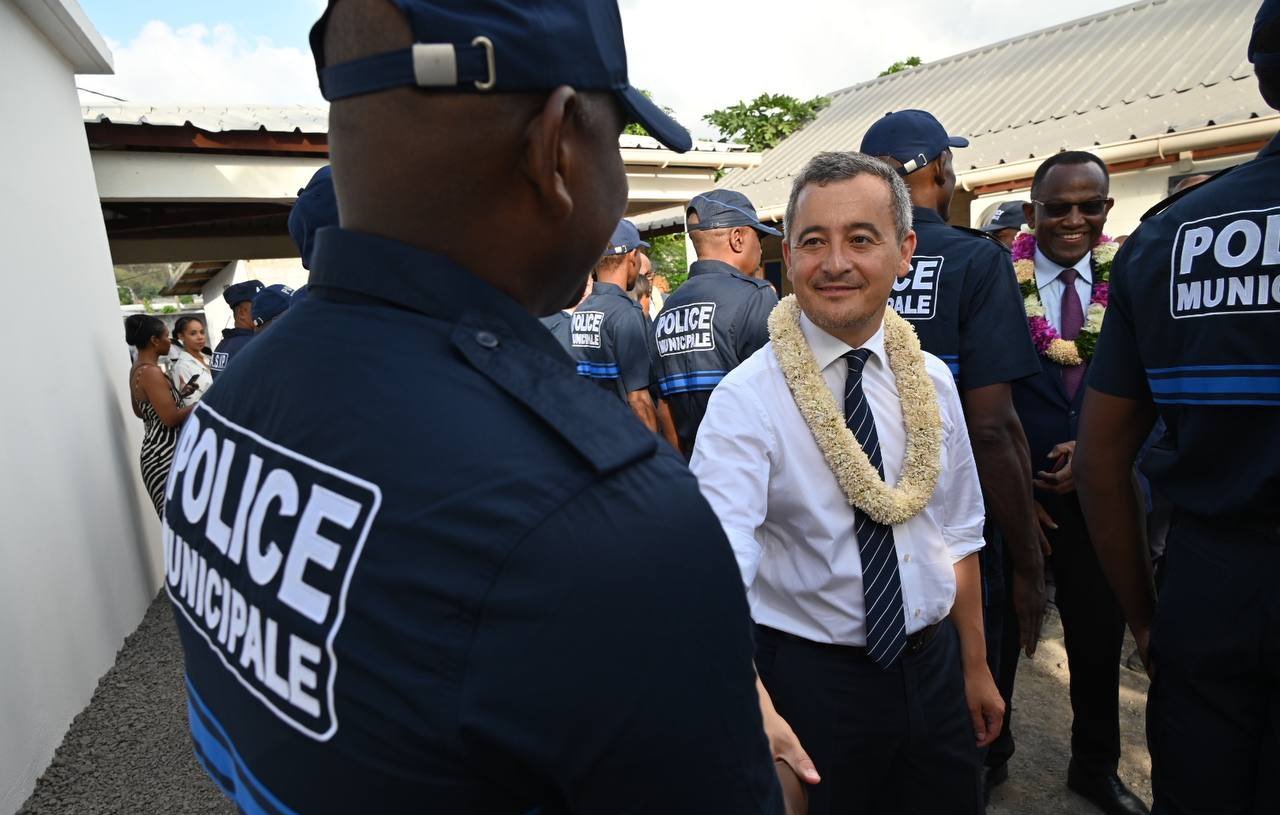 Dans cette image, nous voyons un groupe de policiers municipaux, tous vêtus d'un uniforme bleu avec le mot "POLICE" inscrit sur le dos. Au premier plan, un homme souriant échange une poignée de main avec un policier. Cet homme porte une chemise blanche et une cravate, ainsi qu'un collier de fleurs autour du cou, ce qui suggère un événement formel ou culturel. En arrière-plan, on aperçoit d'autres personnes, dont certaines semblent également habillées de manière soignée. La scène se déroule dans un environnement extérieur, avec des bâtiments aux toits en tôle. L'atmosphère semble accueillante et amicale.
