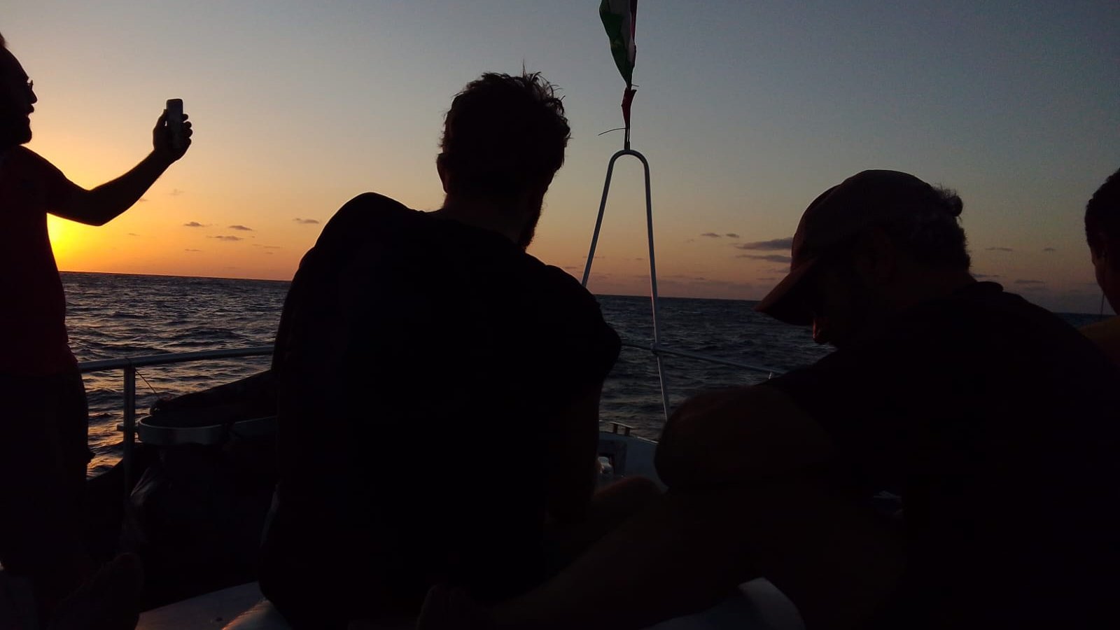 Imaginez une scène sereine sur l'eau, à l'heure où le soleil se couche. Des silhouettes de personnes se découpent contre un fond doré. À gauche, une personne tient un téléphone, capturant le moment, tandis que d'autres sont assises, tournées vers l'horizon. Leurs formes sont sombres, presque spectrales, créant un contraste avec les couleurs chaudes du ciel qui passent du jaune à l'orange et au mauve. La surface de l'eau scintille doucement, reflétant les teintes du crépuscule. L'atmosphère est calme, empreinte de contemplation et d'amitié, tandis que le paysage maritime s'étend à perte de vue.