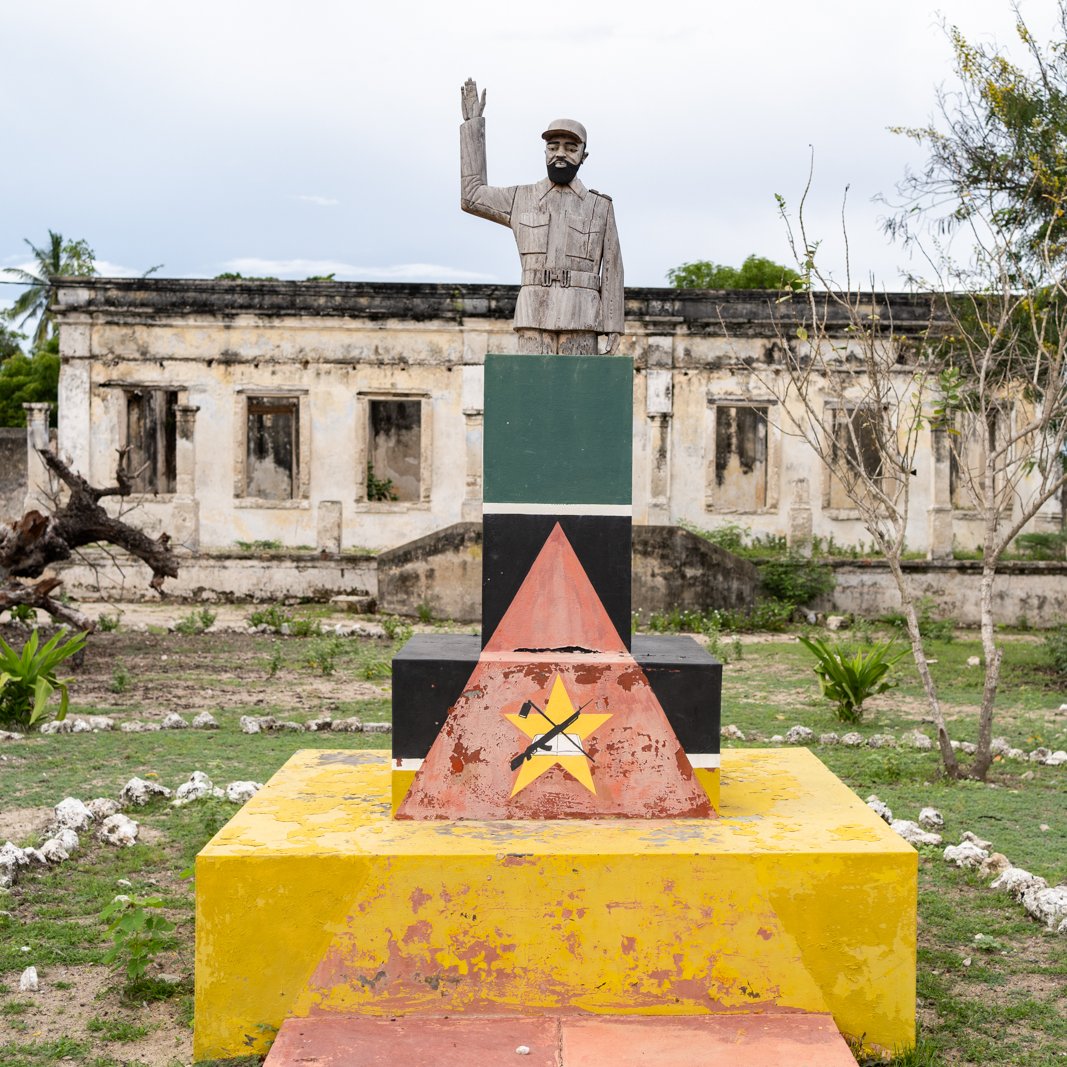 L'image montre un monument composé d'une statue d'un homme saluant, érigée sur une base colorée avec des formes géométriques. La statue est en métal et représente un personnage au regard déterminé. La base est peinte en plusieurs couleurs vives : rouge, jaune, vert et noir, formant des triangles. En arrière-plan, on aperçoit des ruines de bâtiments, avec des murs partiellement effondrés et une végétation verdoyante qui entoure l'ensemble. L'atmosphère est à la fois historique et contemplative, suggérant un lieu chargé de mémoire.