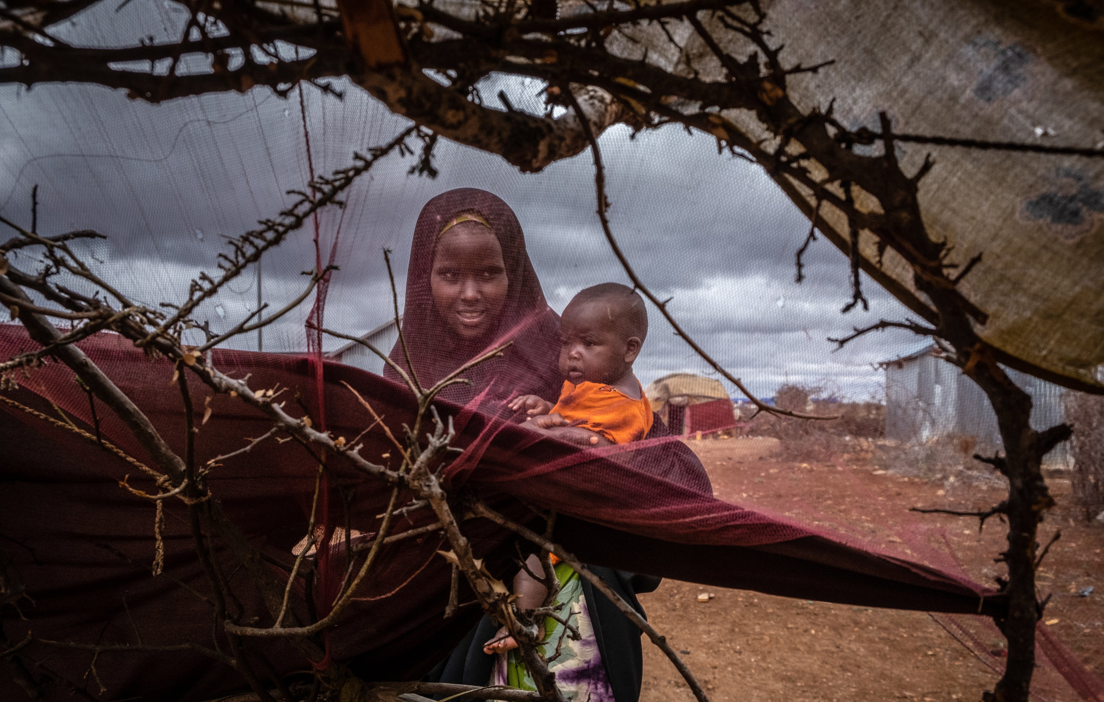 L'image montre une femme et un enfant au sein d'un environnement difficile. La femme, vêtue d'un voile sombre et d'habits traditionnels, tient l'enfant dans ses bras. L'enfant, portant un vêtement orange, semble curieux et regarde autour de lui. Ils sont entourés de branches et de tissus, ce qui forme une sorte d'abri rudimentaire. Le sol est poussiéreux et l'atmosphère est chargée d'une lumière diffuse, suggérant un ciel nuageux. Cette scène évoque la résilience et le lien fort entre la mère et son enfant dans un contexte de précarité.