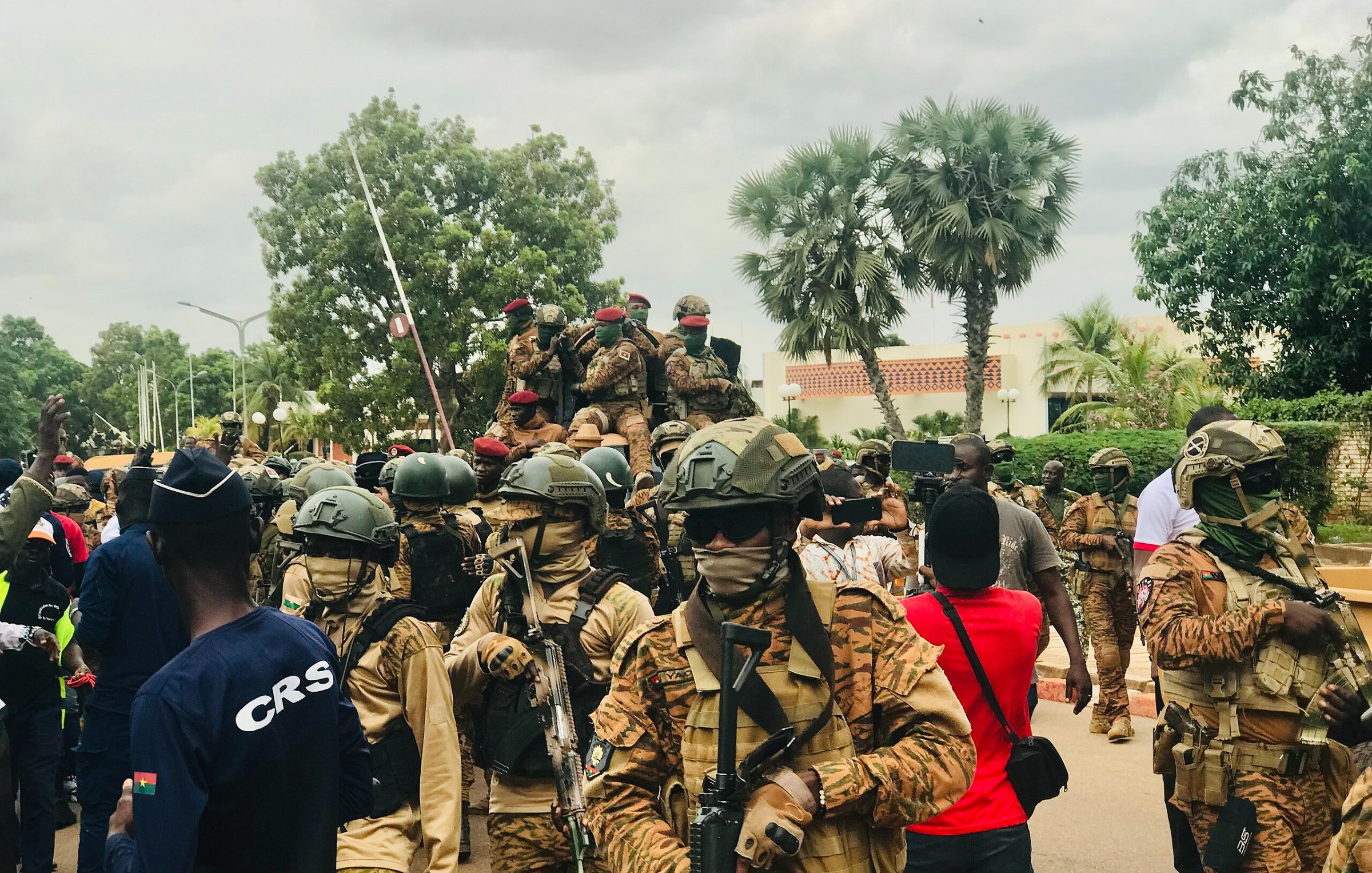 L'image montre une scène animée en plein air, probablement lors d'un événement public ou d'une manifestation. Au premier plan, plusieurs soldats sont regroupés, portant des uniformes militaires et des casques. Ils sont armés et semblent vigilants. L'un d'eux est sur un véhicule militaire, tandis que d'autres avancent sur la route. En arrière-plan, des arbres et des bâtiments peuvent être aperçus, et une foule de citoyens se trouve à proximité, certains portant des vêtements colorés. L'atmosphère peut sembler tendue, surtout en raison de la présence militaire. Le ciel est nuageux, ajoutant une certaine gravité à la scène.