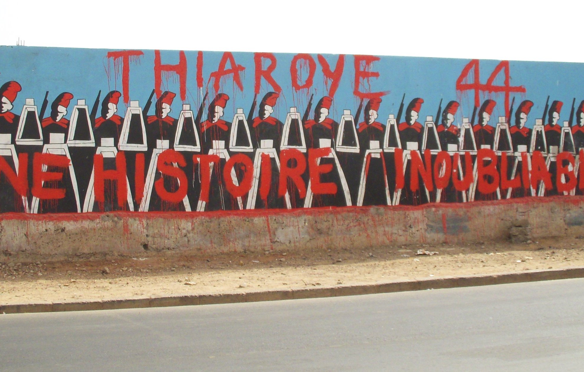 L'image montre une grande fresque murale peinte sur un mur. Au premier plan, on voit une rangée de silhouettes représentant des soldats, chacun portant un uniforme noir et un casque. Ils sont alignés de manière uniforme, les bras le long du corps. En arrière-plan, le mur est peint en bleu, ce qui fait ressortir les personnages. En haut, on peut lire en grandes lettres rouges le texte "THIAROYE 44", et en dessous, "UNE HISTOIRE INOUBLIABLE". Cette œuvre évoque une mémoire historique importante, probablement liée à des événements marquants qui ont eu lieu à Thiaroye, une ville du Sénégal. L'ensemble de l'image crée une ambiance à la fois sérieuse et respectueuse, honorant un passé collectif.