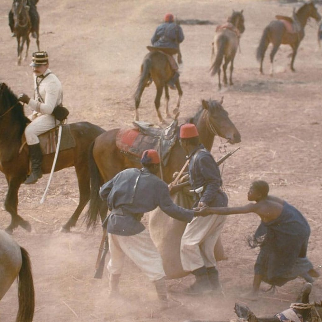 L'image présente une scène historique en plein air, où plusieurs personnages sont engagés dans une interaction dynamique. Au centre, un homme vêtu d'un uniforme militaire blanc est assis sur un cheval, observant la scène. Deux autres hommes, probablement des soldats, sont en train de se battre ou de grappler au sol, mettant en évidence un moment de tension. En arrière-plan, plusieurs autres chevaux et hommes se déplacent dans un environnement poussiéreux, suggérant une ambiance mouvementée et chaotique, typique d'un camp militaire. Le terrain est sec, peut-être de la terre battue, ce qui ajoute à l'atmosphère rustique et historique de la scène.