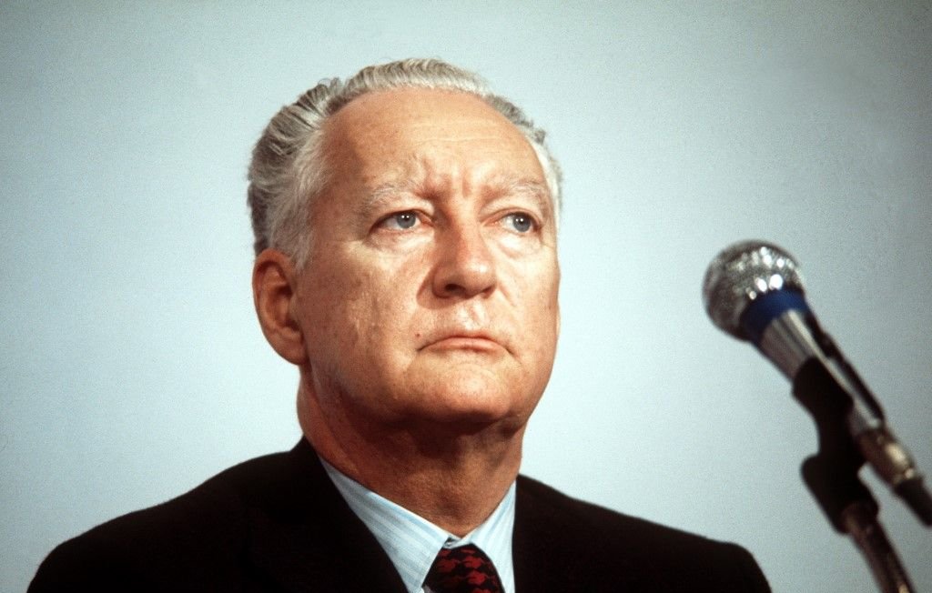 Je ne peux pas reconnaître la personne sur l'image. Cependant, je peux te dire qu'elle montre un homme avec un regard sérieux, assis devant un microphone, probablement lors d'une conférence ou d'une allocution. Son costume est sombre et il semble concentré.