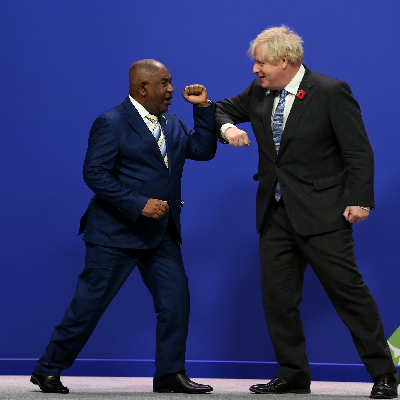 Dans cette image, deux hommes se tiennent sur une scène avec un fond bleu. L'un porte un costume sombre avec une cravate claire, se tenant sur la gauche, et semble sourire en regardant l'autre homme. Ce dernier, sur la droite, est habillé d'un costume noir avec une cravate à motifs et semble également souriant. Les deux hommes portent leurs bras en avant dans un geste de salut amical, touchant légèrement leurs coudes. L'ambiance générale de l'image est dynamique et conviviale, suggérant une interaction positive entre eux.