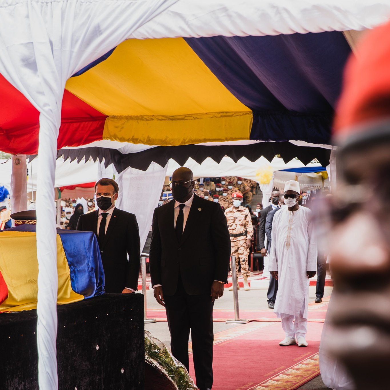 L'image montre une scène d'événement officiel, probablement une cérémonie. Au premier plan, il y a un homme portant des lunettes et un uniforme sombre, dont le visage est flou mais qui semble attentif. En arrière-plan, deux autres hommes, l'un en costume noir et l'autre en tenue traditionnelle blanche, marchent sous un chapiteau aux couleurs vives de jaune, rouge et bleu. Le chapiteau est décoré de tissu et crée une atmosphère formelle. La foule semble nombreuse et attentive, rendant l'ensemble de la scène solennelle et respectueuse. L'éclairage semble naturel, suggérant peut-être une belle journée ensoleillée.
