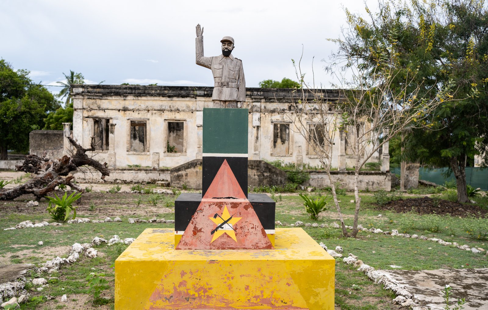 L'image montre un monument composé d'une statue d'un homme saluant, érigée sur une base colorée avec des formes géométriques. La statue est en métal et représente un personnage au regard déterminé. La base est peinte en plusieurs couleurs vives : rouge, jaune, vert et noir, formant des triangles. En arrière-plan, on aperçoit des ruines de bâtiments, avec des murs partiellement effondrés et une végétation verdoyante qui entoure l'ensemble. L'atmosphère est à la fois historique et contemplative, suggérant un lieu chargé de mémoire.