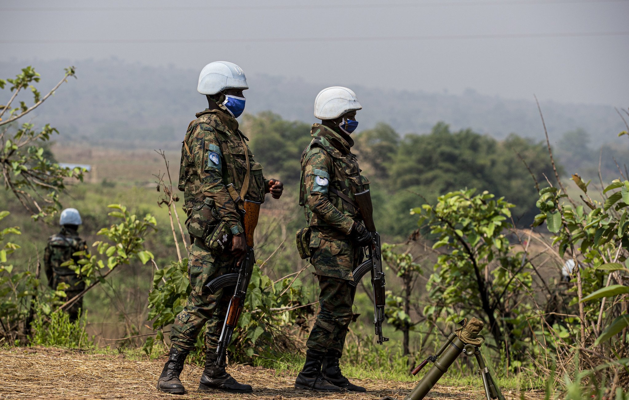 L'image montre deux militaires en uniforme, portant des casques blancs et des masques. Ils se tiennent debout sur un terrain légèrement vallonné, entourés de verdure, avec des plantes et des arbres en arrière-plan. Les soldats tiennent des armes et semblent vigilants, observant leur environnement. L'atmosphère est sérieuse, évoquant un contexte de mission ou de sécurité. Le ciel est clair, et l'ambiance générale peut donner une impression de calme malgré la présence militaire.